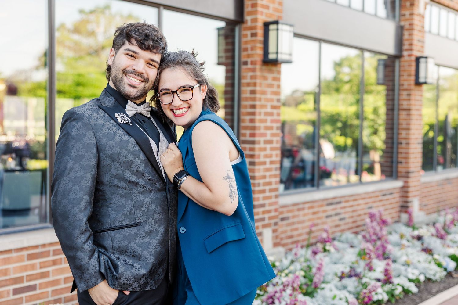 A happy couple stands together smiling in front of a brick building with pink flowers blooming nearby.