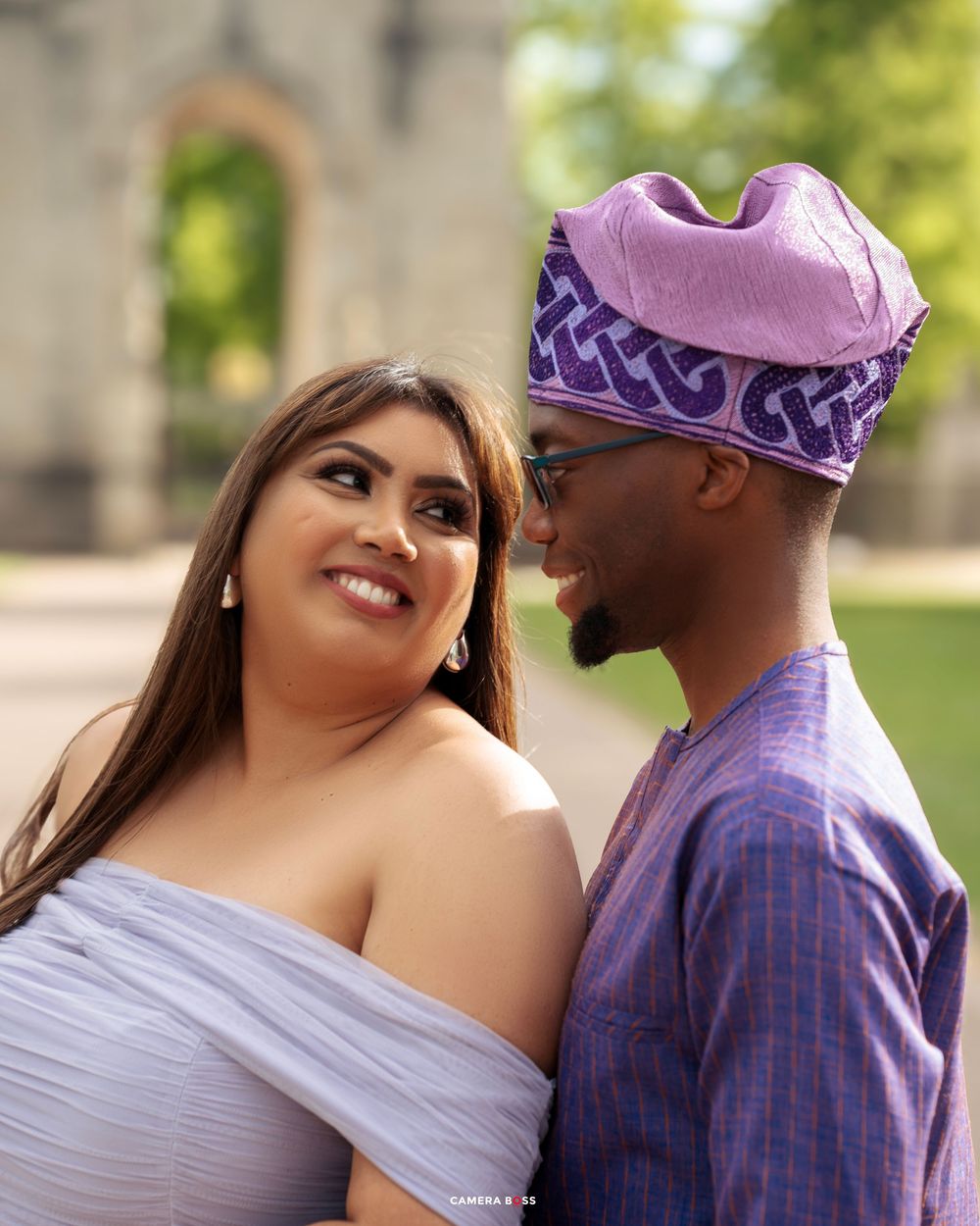 A couple shares a romantic moment outdoors, with one wearing a purple patterned traditional hat and the other in a lavender dress.