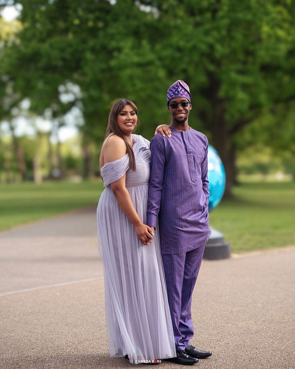 A couple poses together outdoors in coordinating purple outfits on a paved path surrounded by green grass.