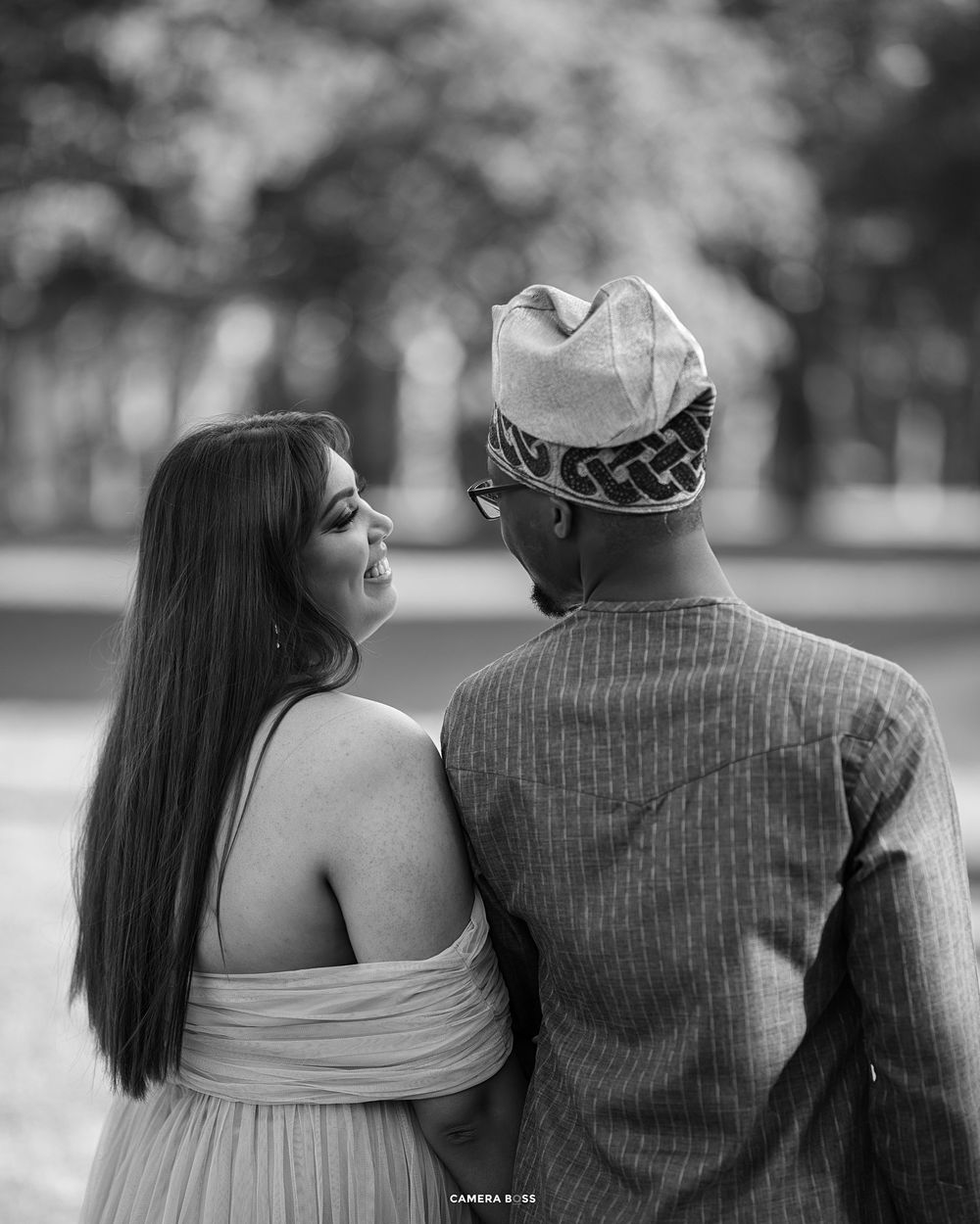 Black and white portrait of a couple in outdoor setting with woman in dress looking back at partner wearing traditional cap.