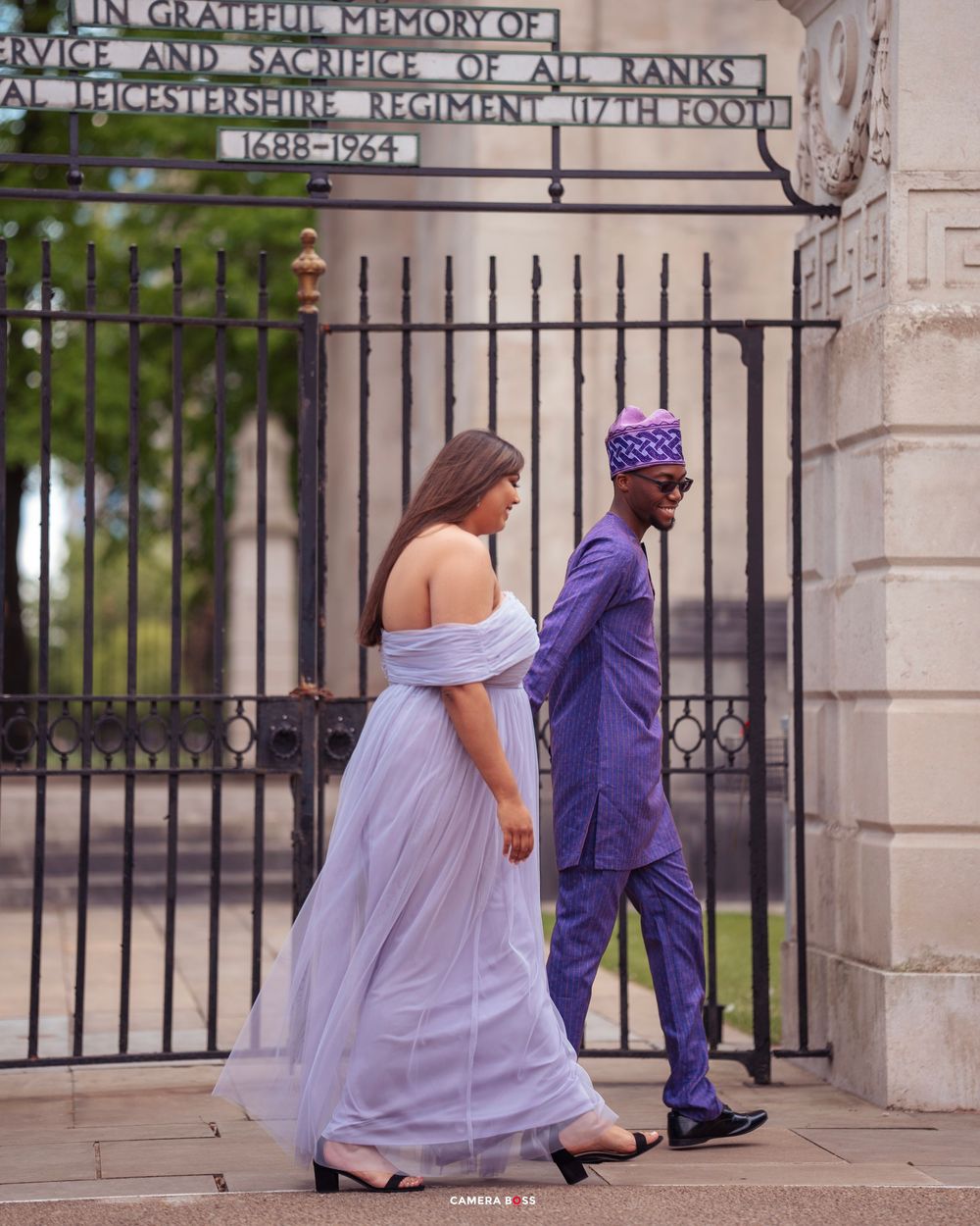 A couple in contrasting purple and lilac outfits walk through ornate iron gates at a historic building entrance.