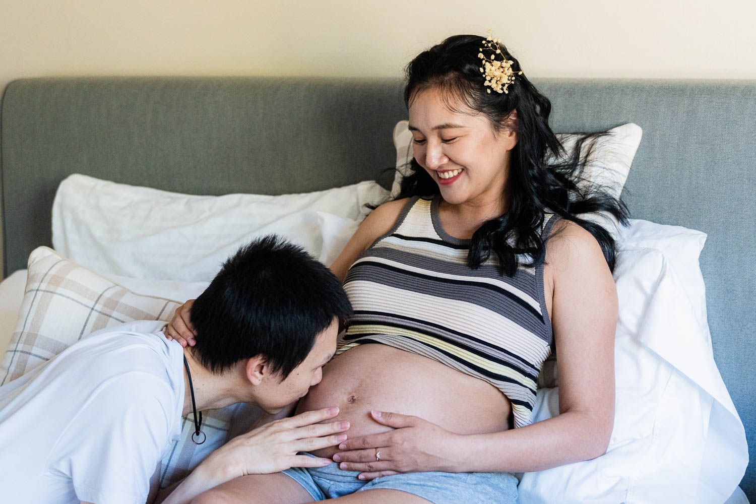 Pregnant partner resting on bed while partner kisses baby bump through striped maternity shirt.
