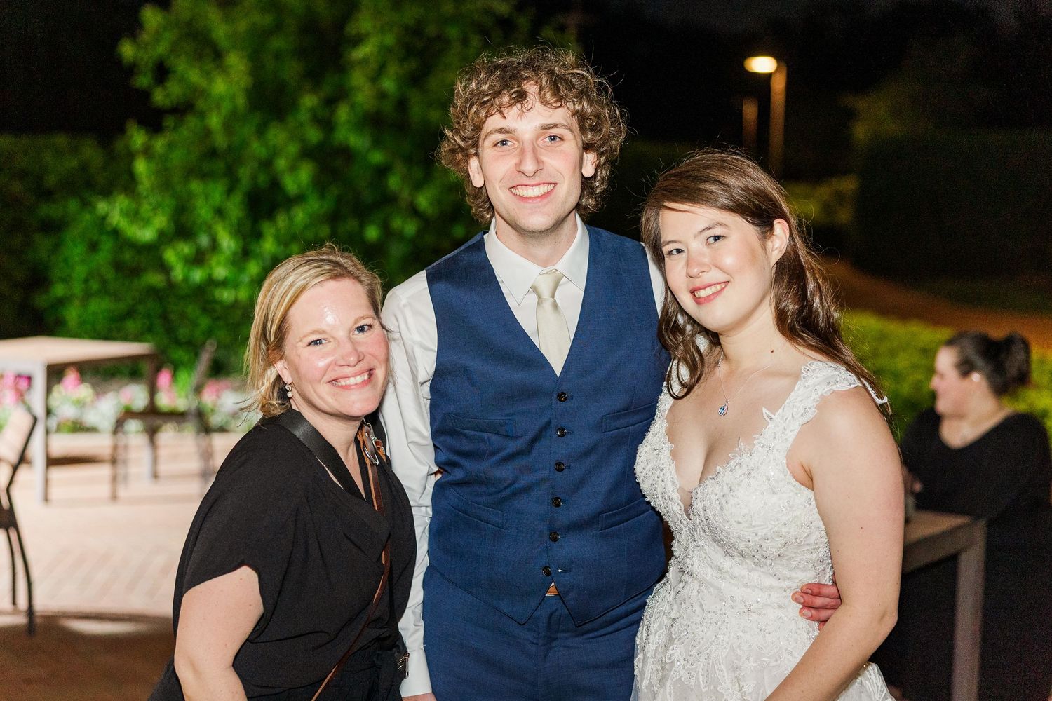 A bride in a white dress and groom in a blue vest smile with a guest at an outdoor evening wedding reception.