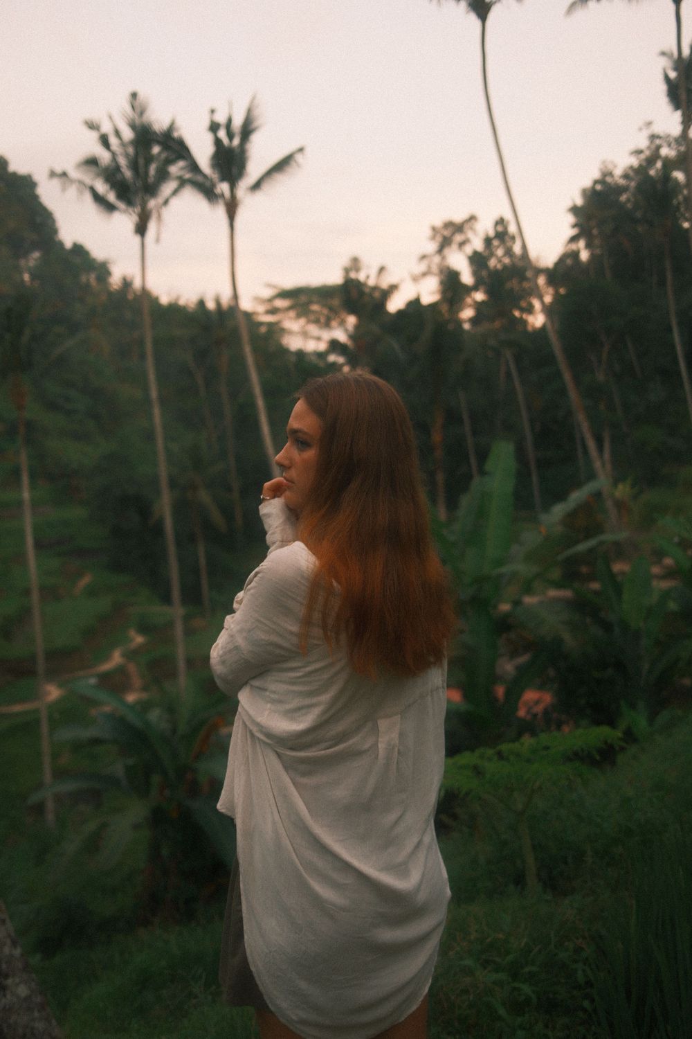 Woman in flowing white dress poses against backdrop of tropical palm trees and terraced hills at sunset.