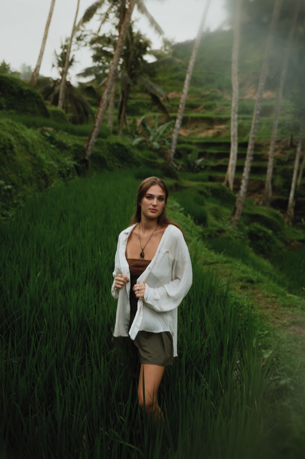 Person in white shirt and brown top amid lush rice fields and palm trees.