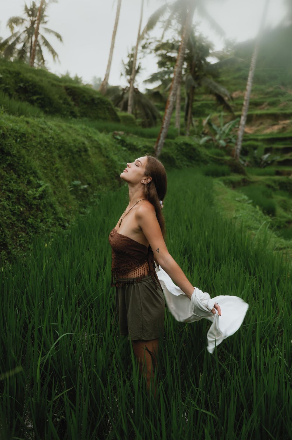 Woman in flowing dress poses dramatically in lush Balinese rice terrace landscape.