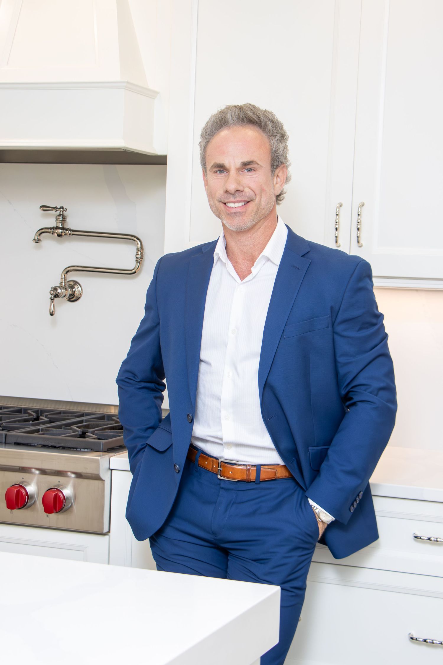 Professional in a navy blue suit and white shirt stands confidently in a modern white kitchen with stainless steel appliances.