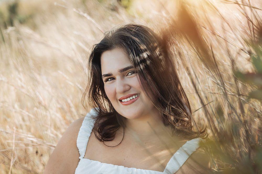 Smiling young woman in white dress standing among tall wheat grass in golden sunlight.