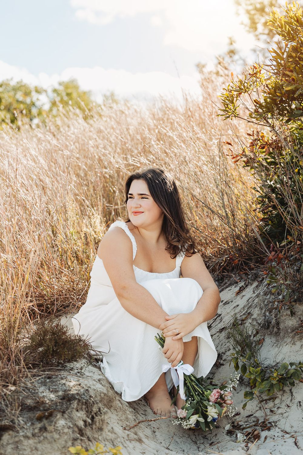 Four dreamy outdoor portraits of someone wearing a white dress sitting on coastal sand dunes at golden hour.