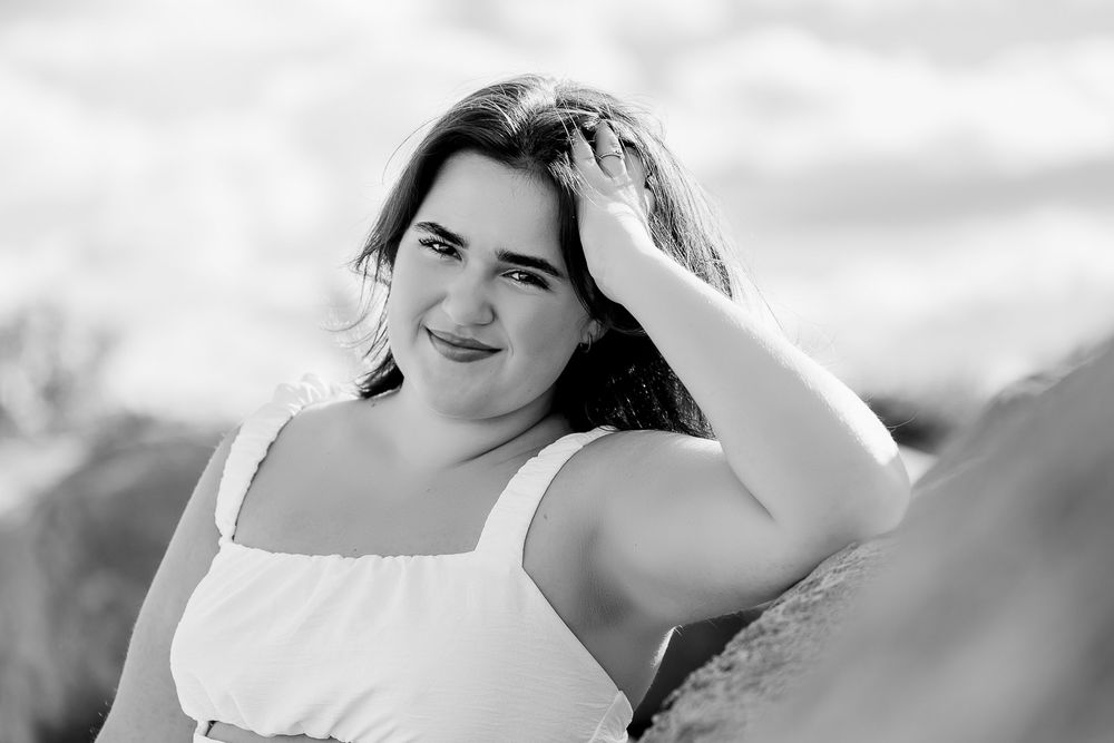 Black and white portrait of a woman in a white top running her hand through her hair against a cloudy sky.