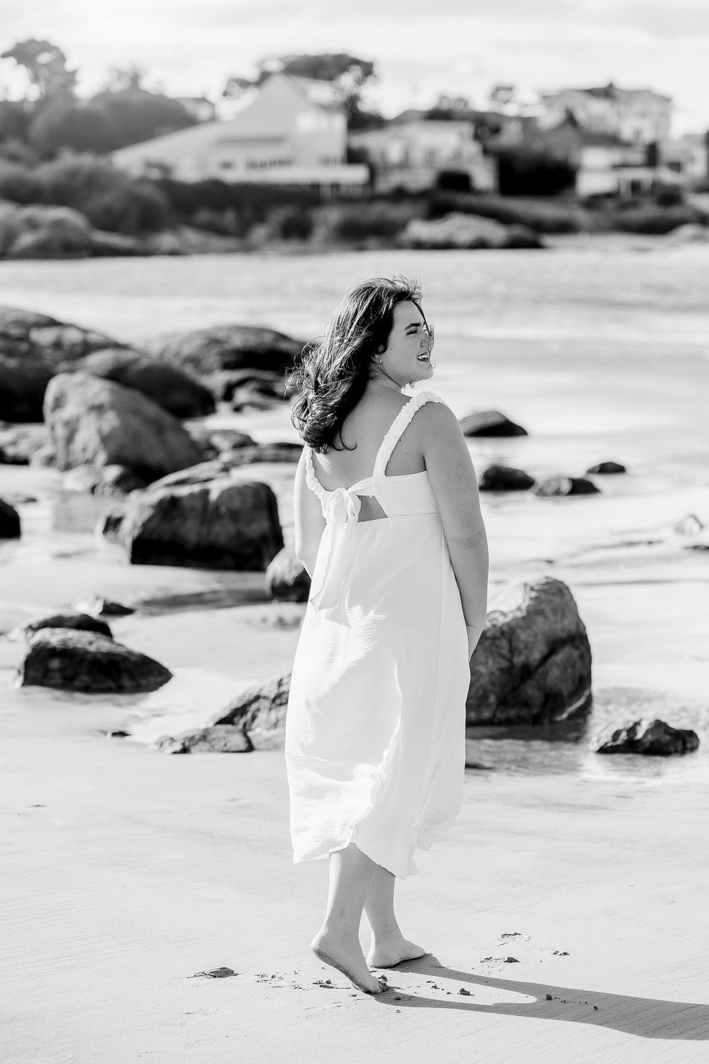 A figure in a flowing white dress walks gracefully along a rocky beach shoreline in black and white photography.