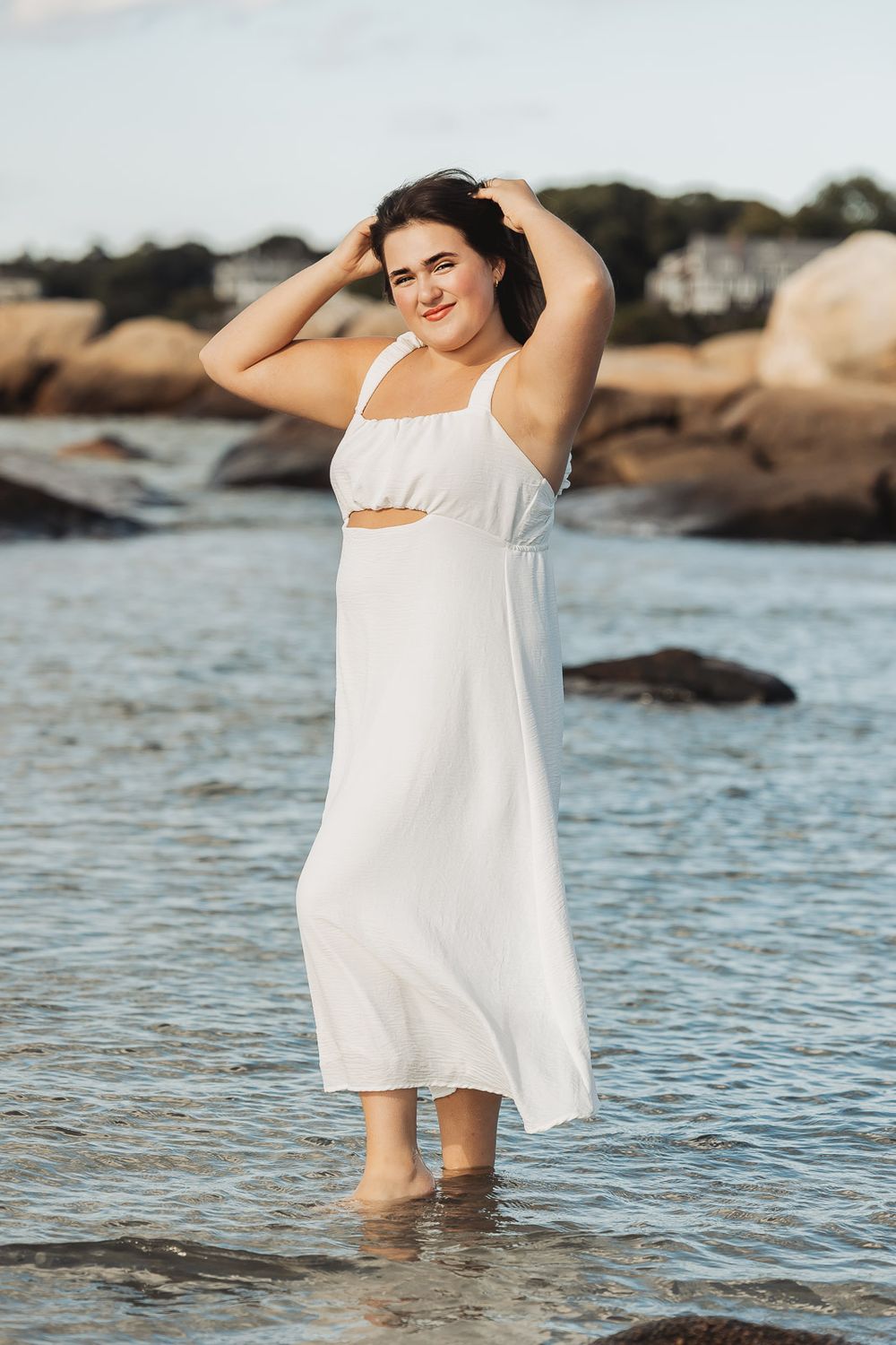Someone wearing a flowing white dress walks through shallow ocean waters near rocky coastline at sunset.