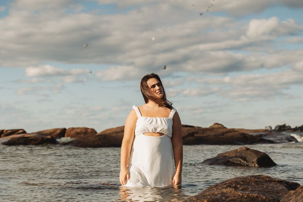 Person in a white sundress standing in shallow ocean water among rocky outcrops under a cloudy sky.