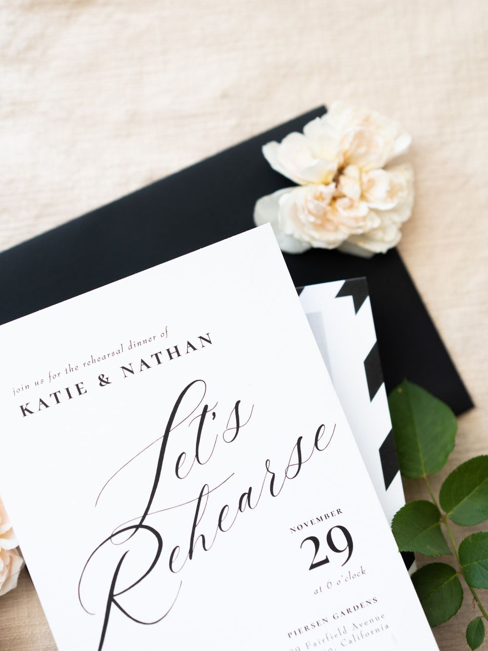 Wedding invitation with elegant script text showing 'Let's Rehearse' on a black background with white roses and greenery.