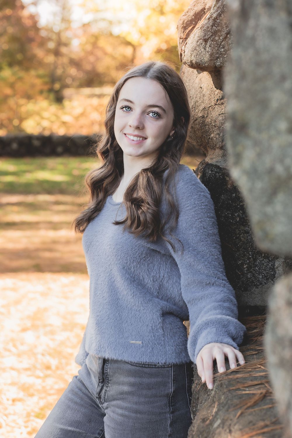 Person in a gray sweater leaning against a stone wall in an autumn setting with golden foliage.