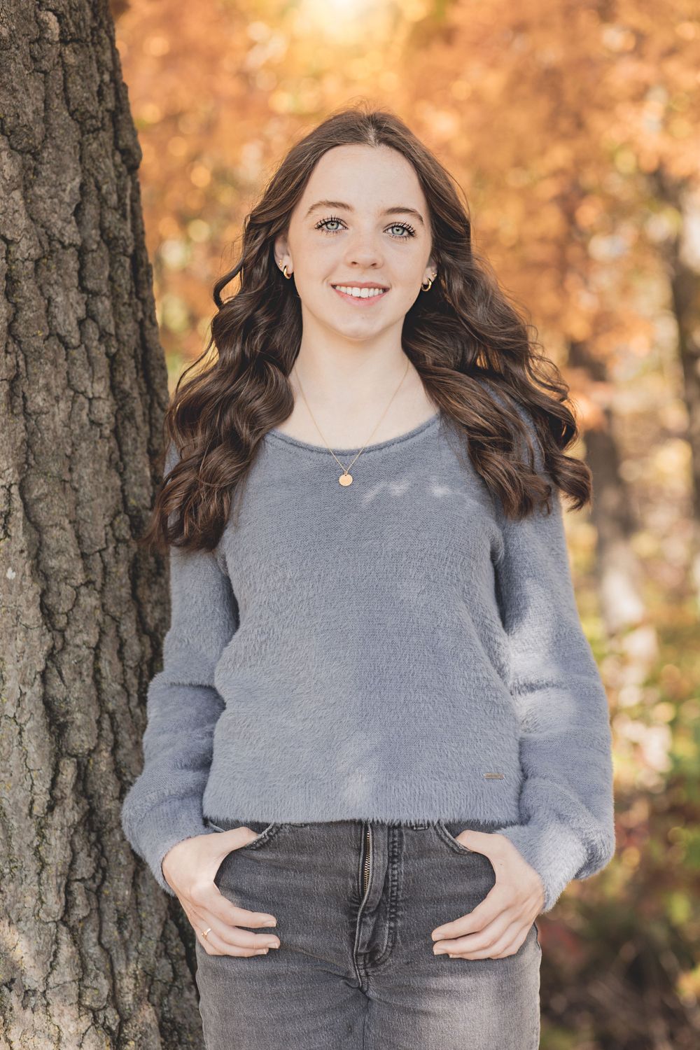 Person in grey sweater poses against tree trunk with autumn colors in background.