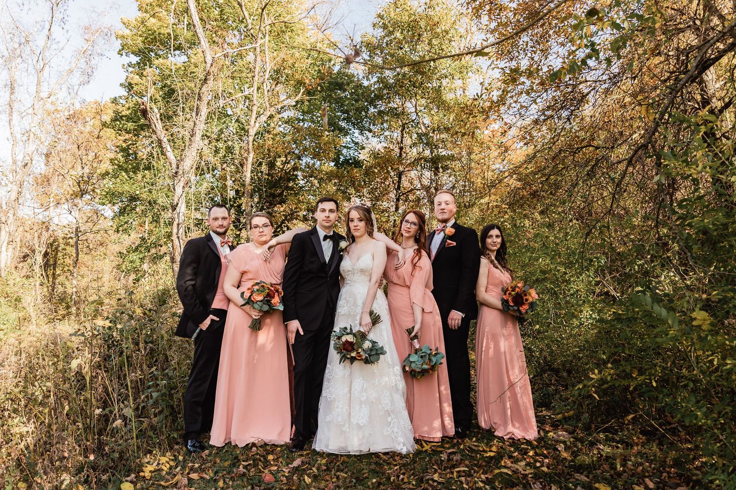 A wedding party poses together in a scenic wooded area on a sunny autumn day while wearing pink and white dresses.