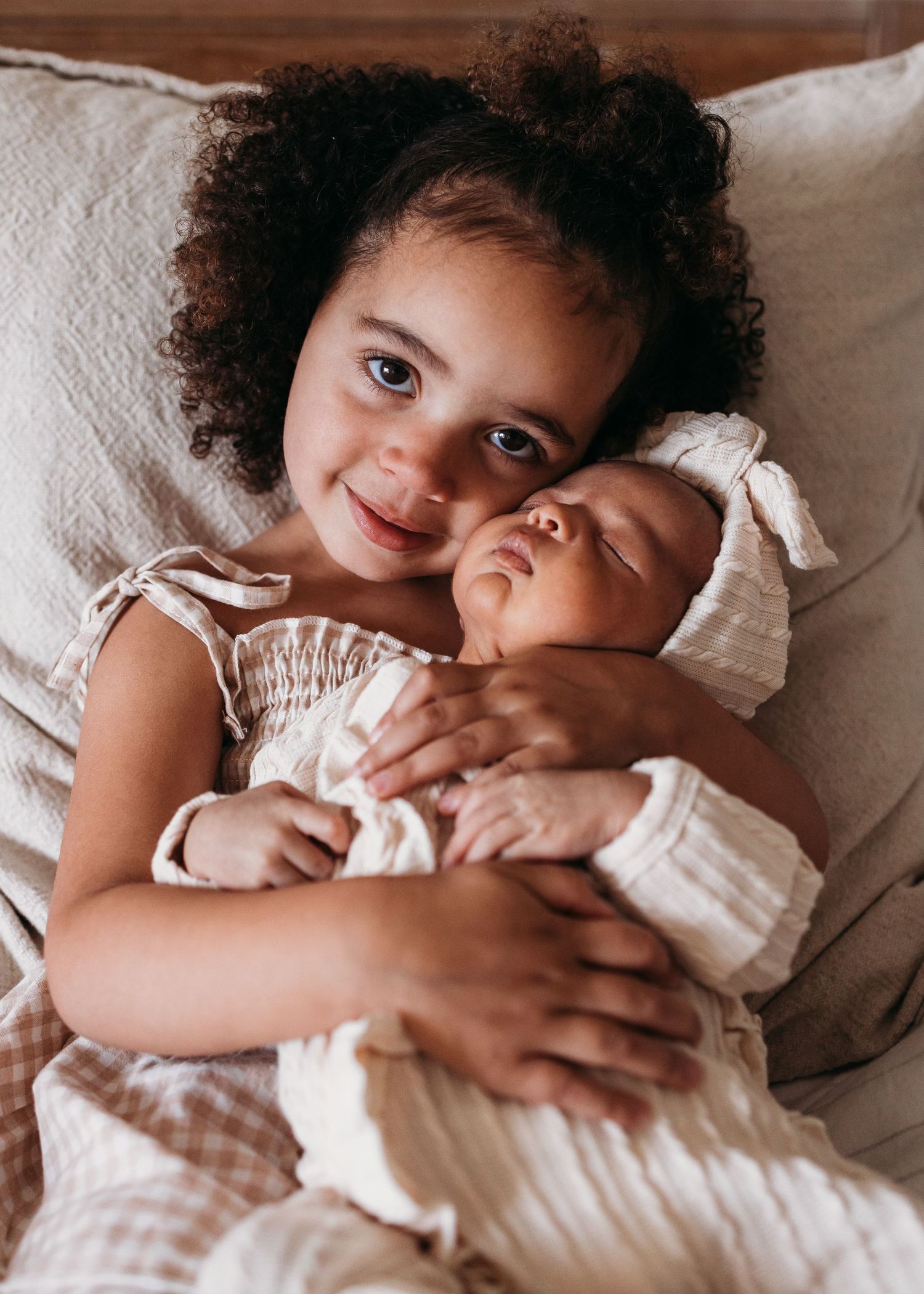 Two young children in white outfits share a tender embrace while lying on white bedding, creating a sweet moment.