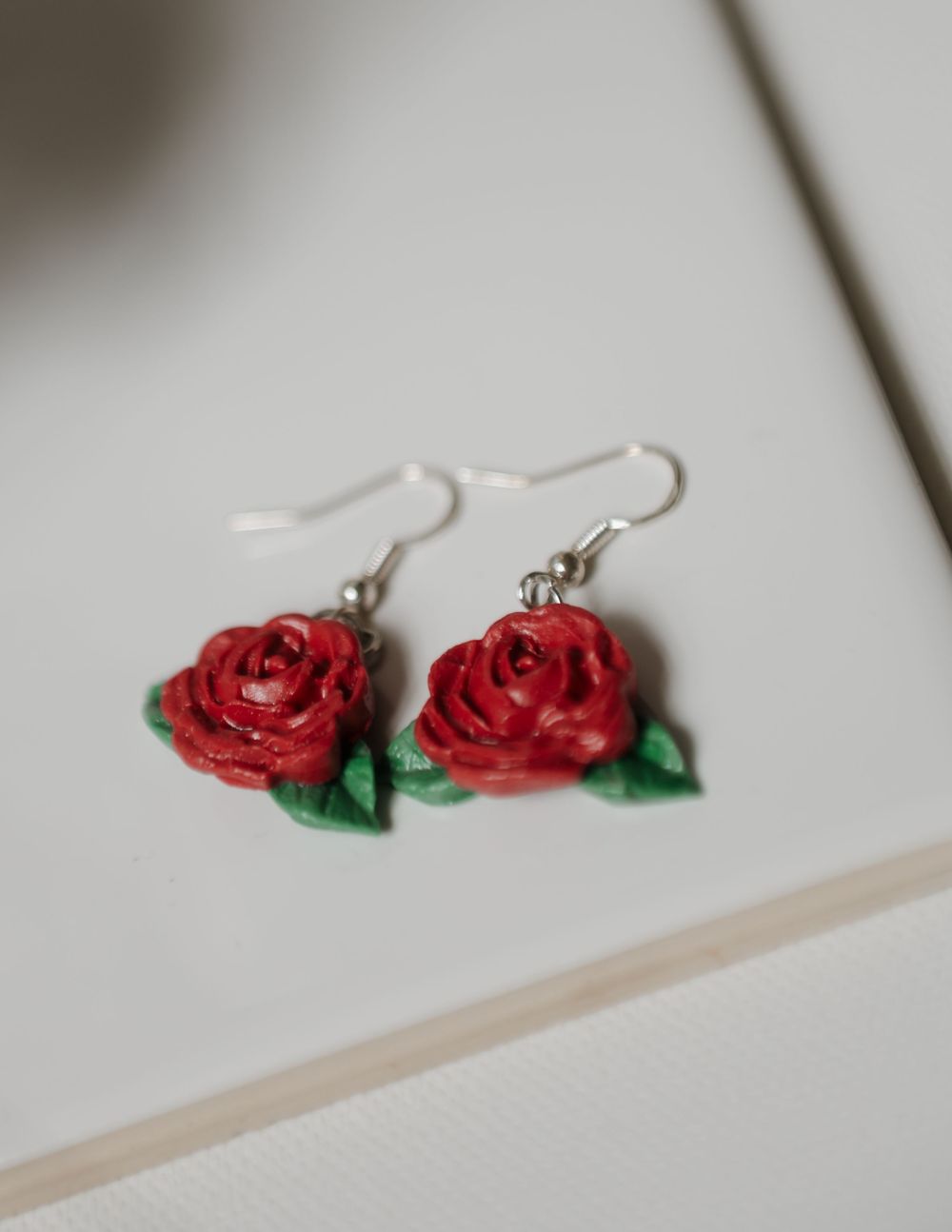 A pair of red rose earrings with green leaves on silver hooks displayed on a white background.