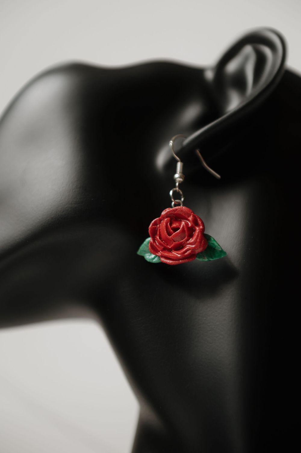 Delicate red rose earring displayed on a black mannequin head profile against a light gray background.