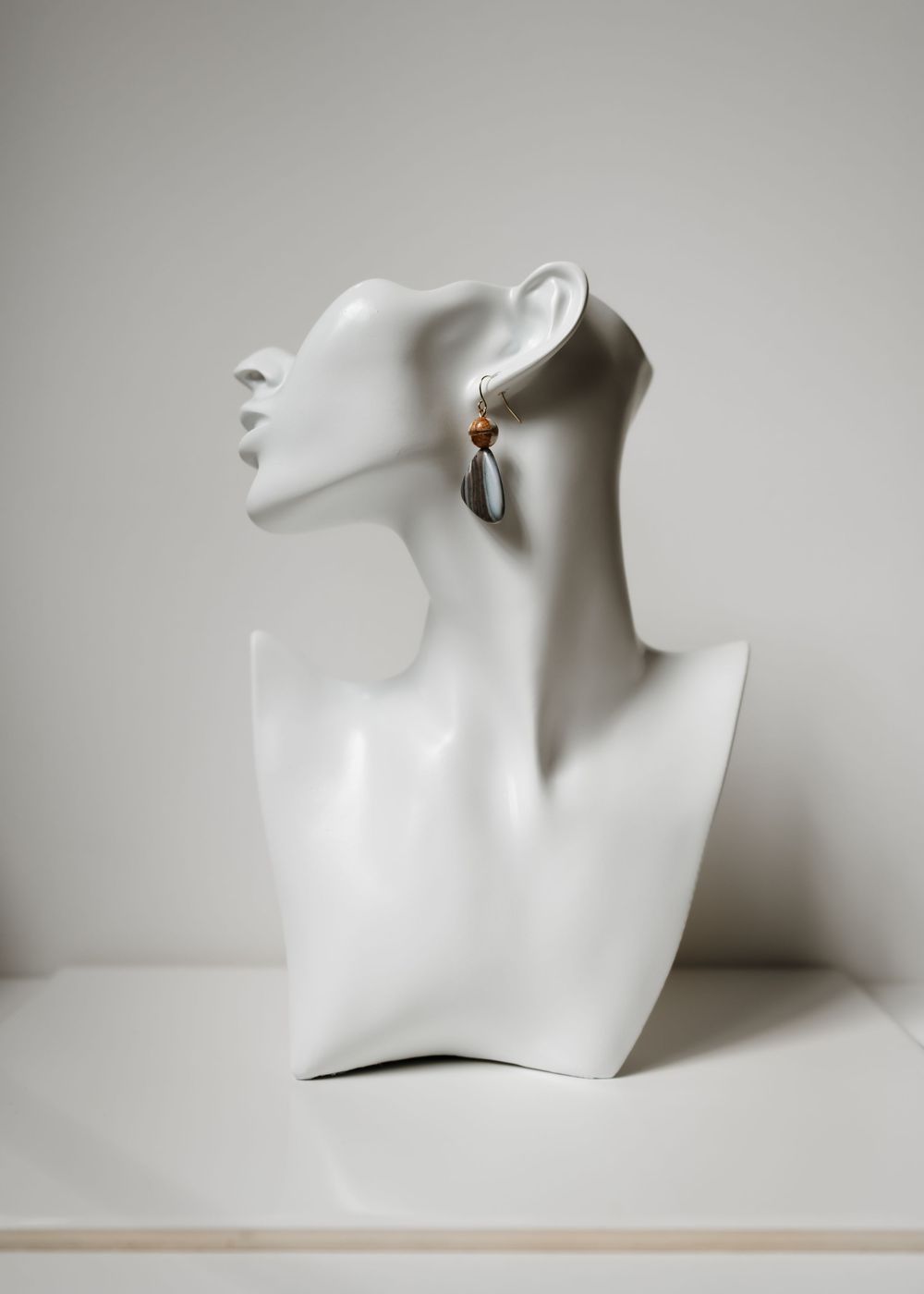 A white fashion mannequin bust with an elegant neck and profile displays a dangling earring against a neutral grey background.