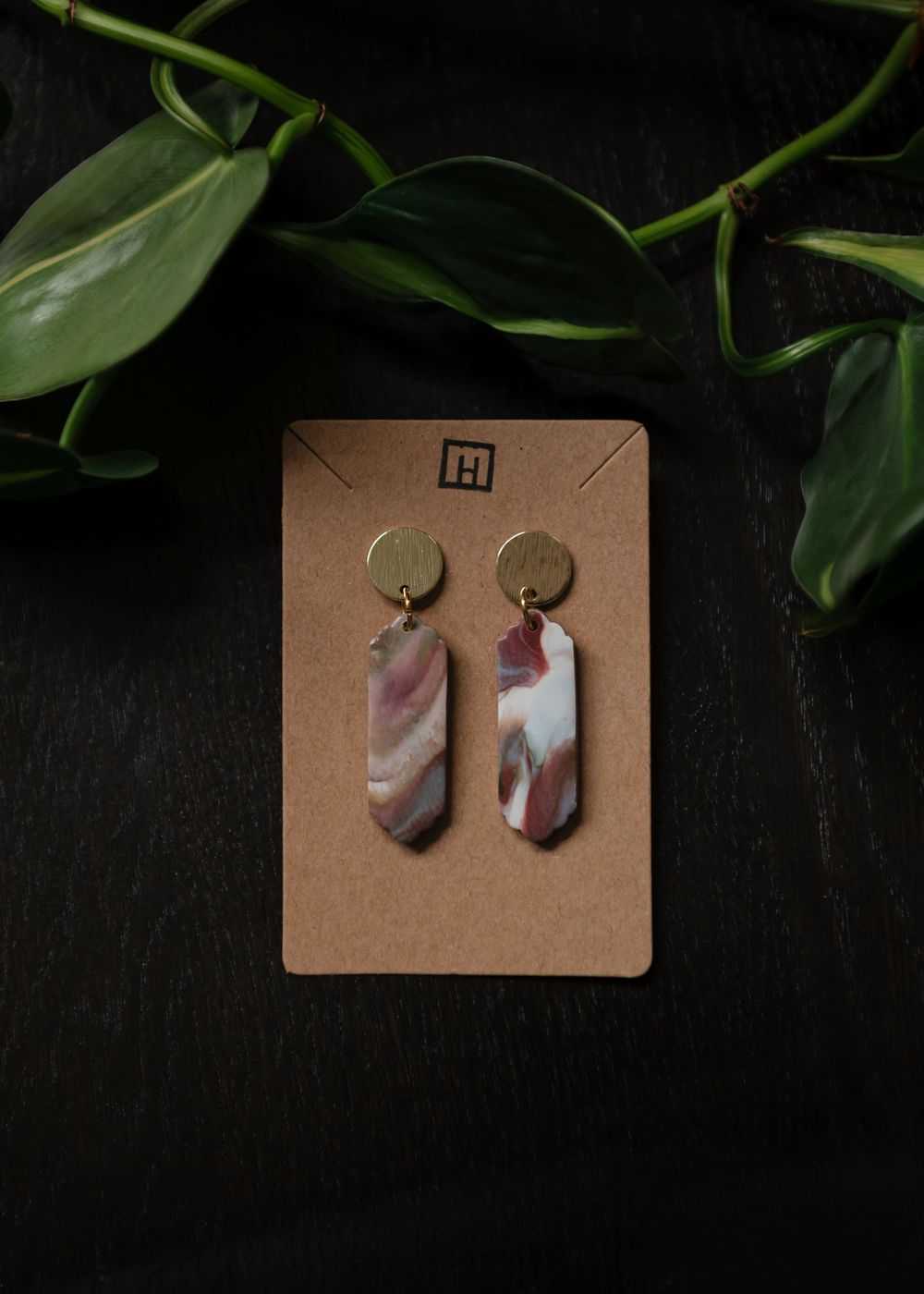 Hand-crafted polymer clay earrings in marbled brown and white tones displayed on a kraft paper card with green foliage.