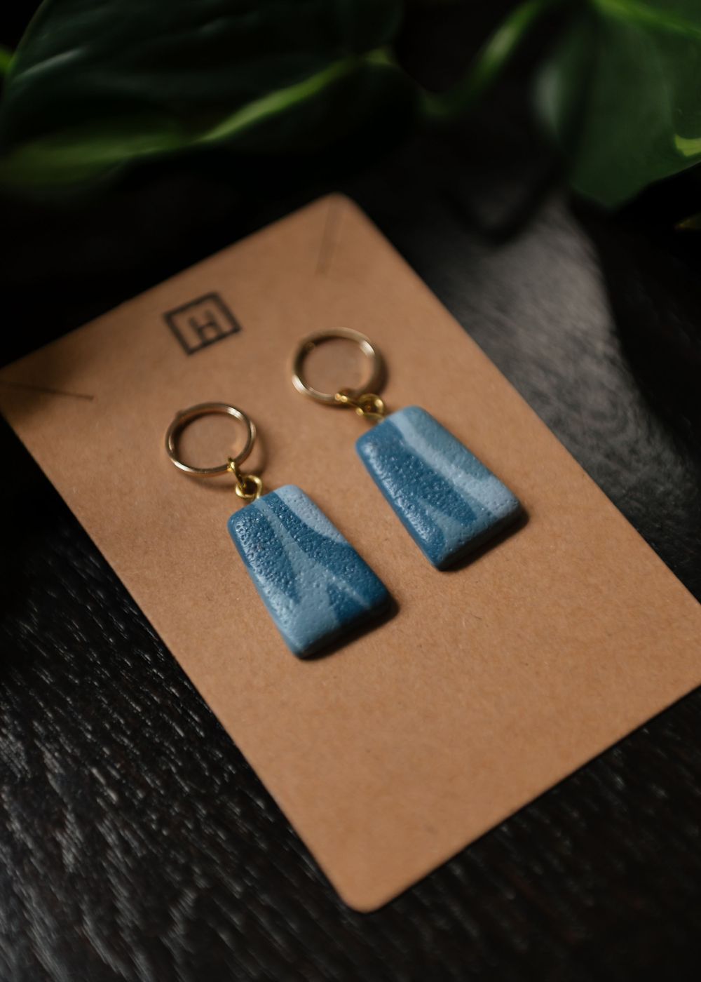 Handmade blue clay trapezoid earrings with gold hoops displayed on brown craft paper against dark surface.