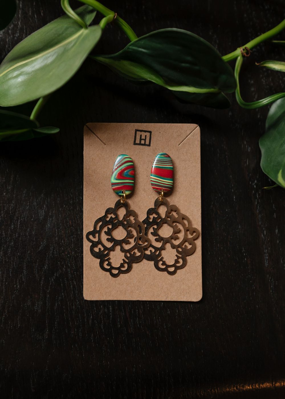 Handmade striped earrings with red and green patterns on wooden cutouts displayed on brown cardboard with green leaves.