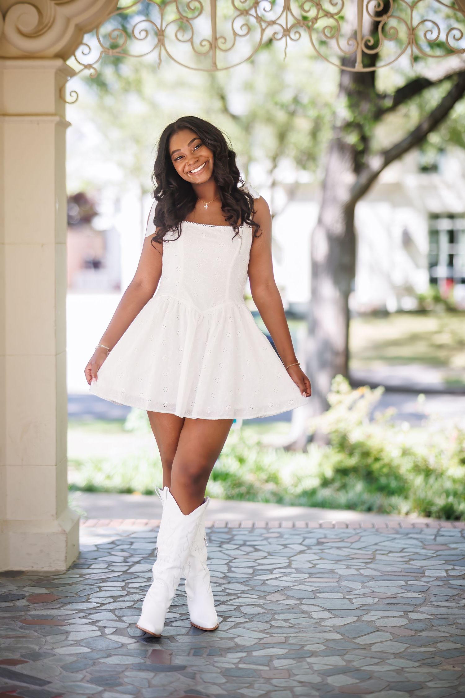 Young woman in a white summer dress and knee-high white boots poses under an ornate archway on a cobblestone path for her graduation photos