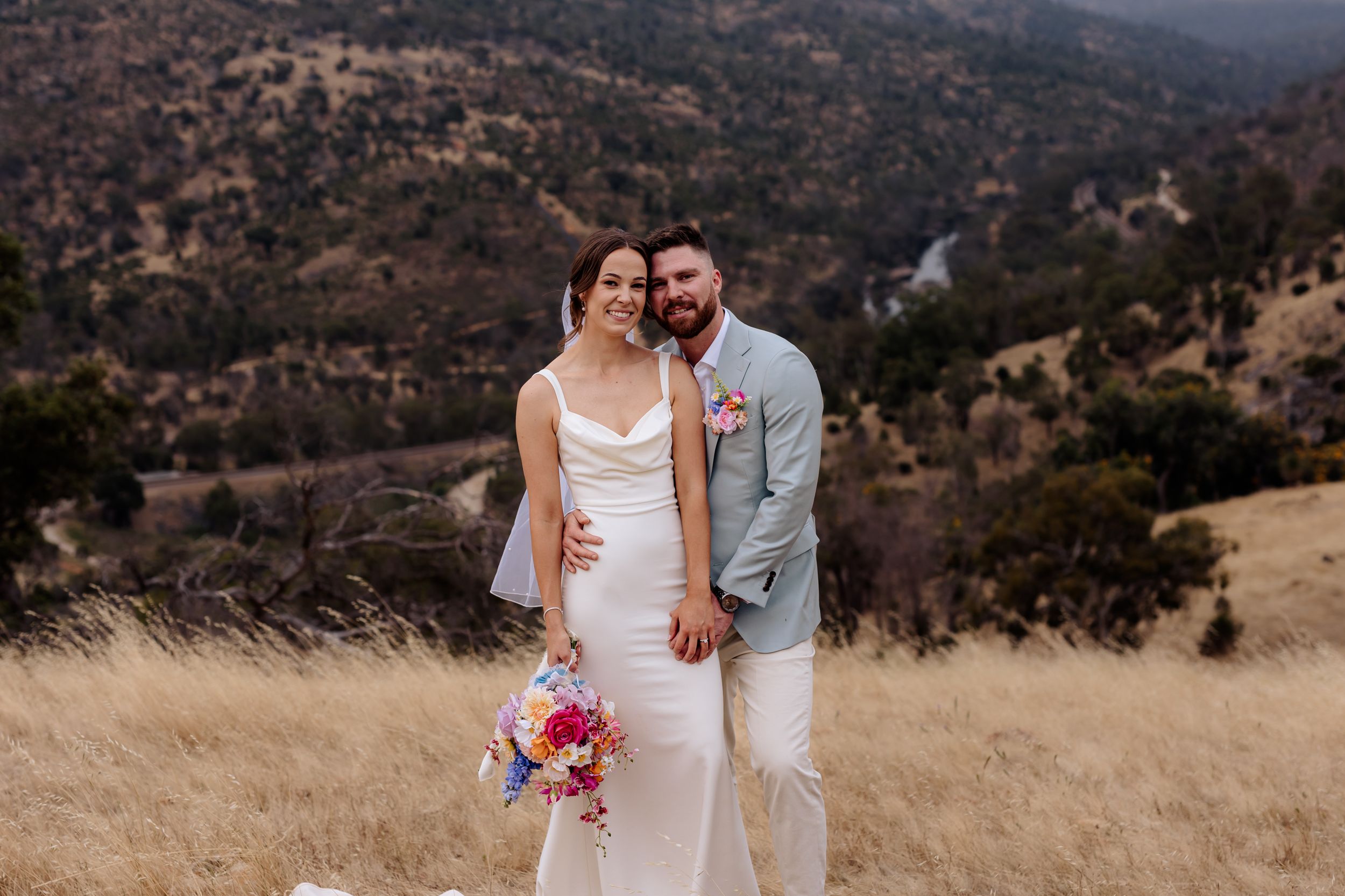 A couple stands together in a light gray suit and white dress holding a colorful bouquet on a golden hillside landscape.