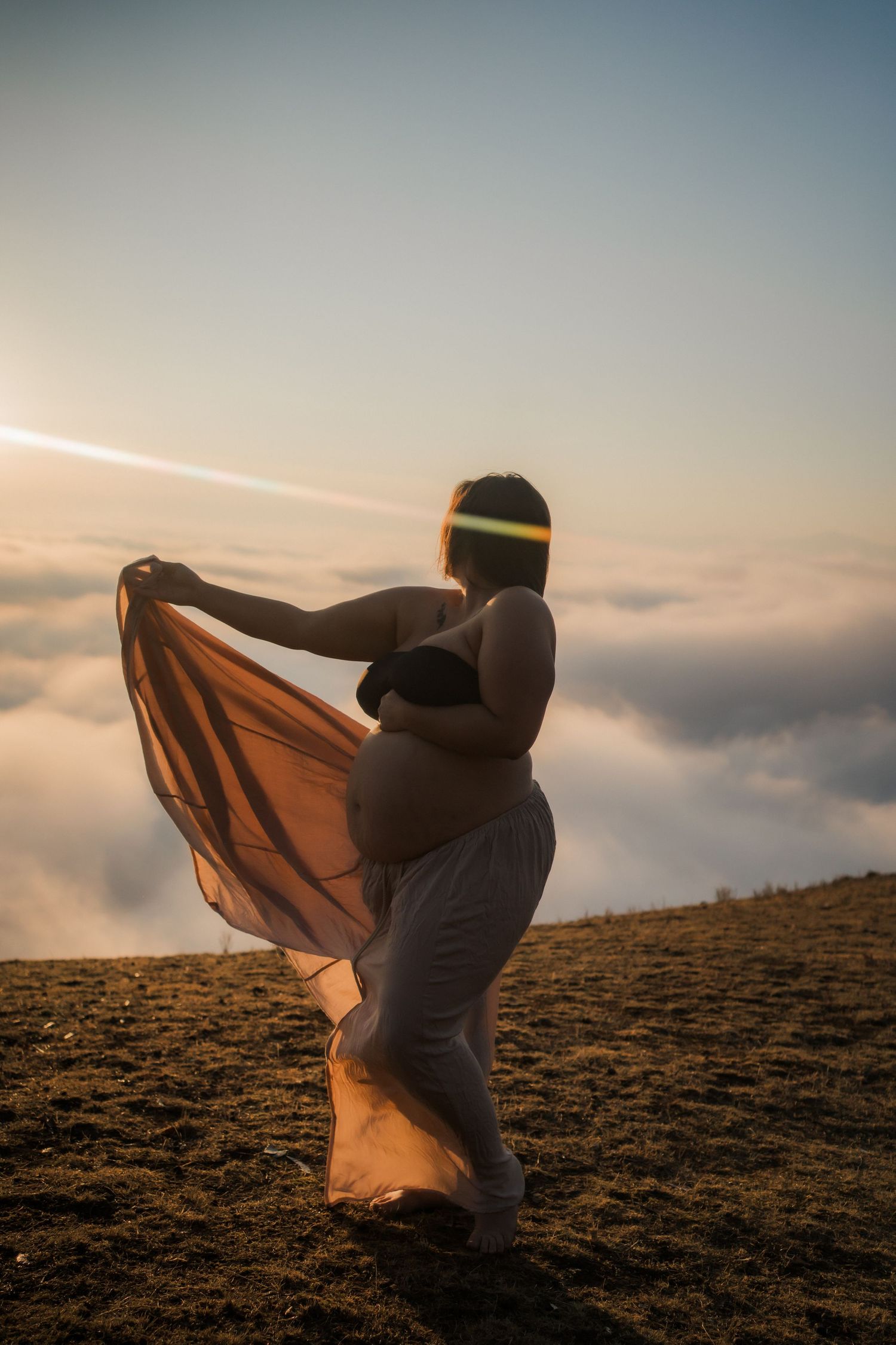 Silhouette of pregnant mum in flowing fabric standing on hilltop against cloudy sunset sky in golden light in Mansfield, Vic High Country