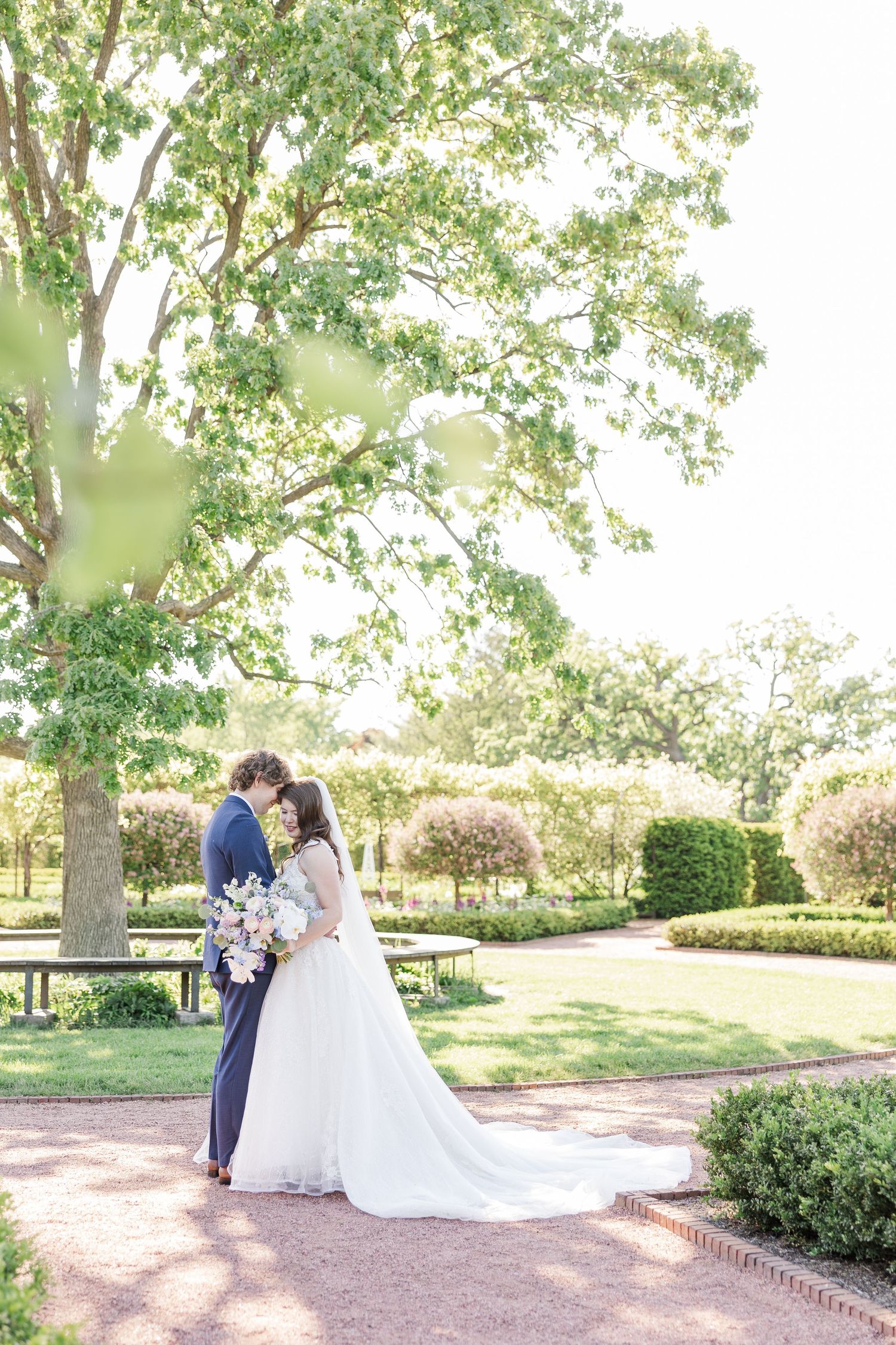 Wedding couple embraces in sunlit garden setting with lush trees and manicured landscaping on a bright summer day.