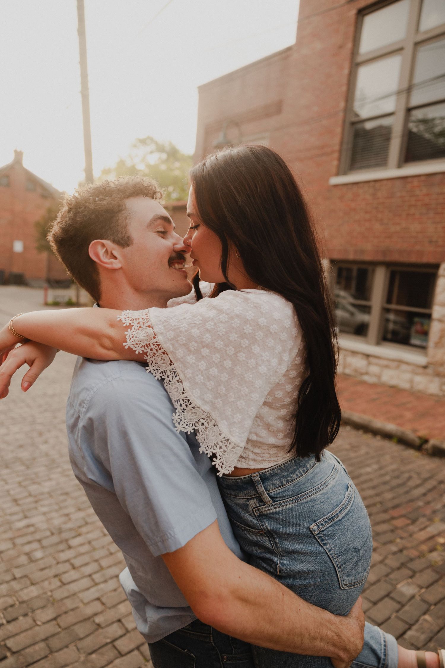 A romantic couple embracing on a brick sidewalk at sunset near historic buildings.