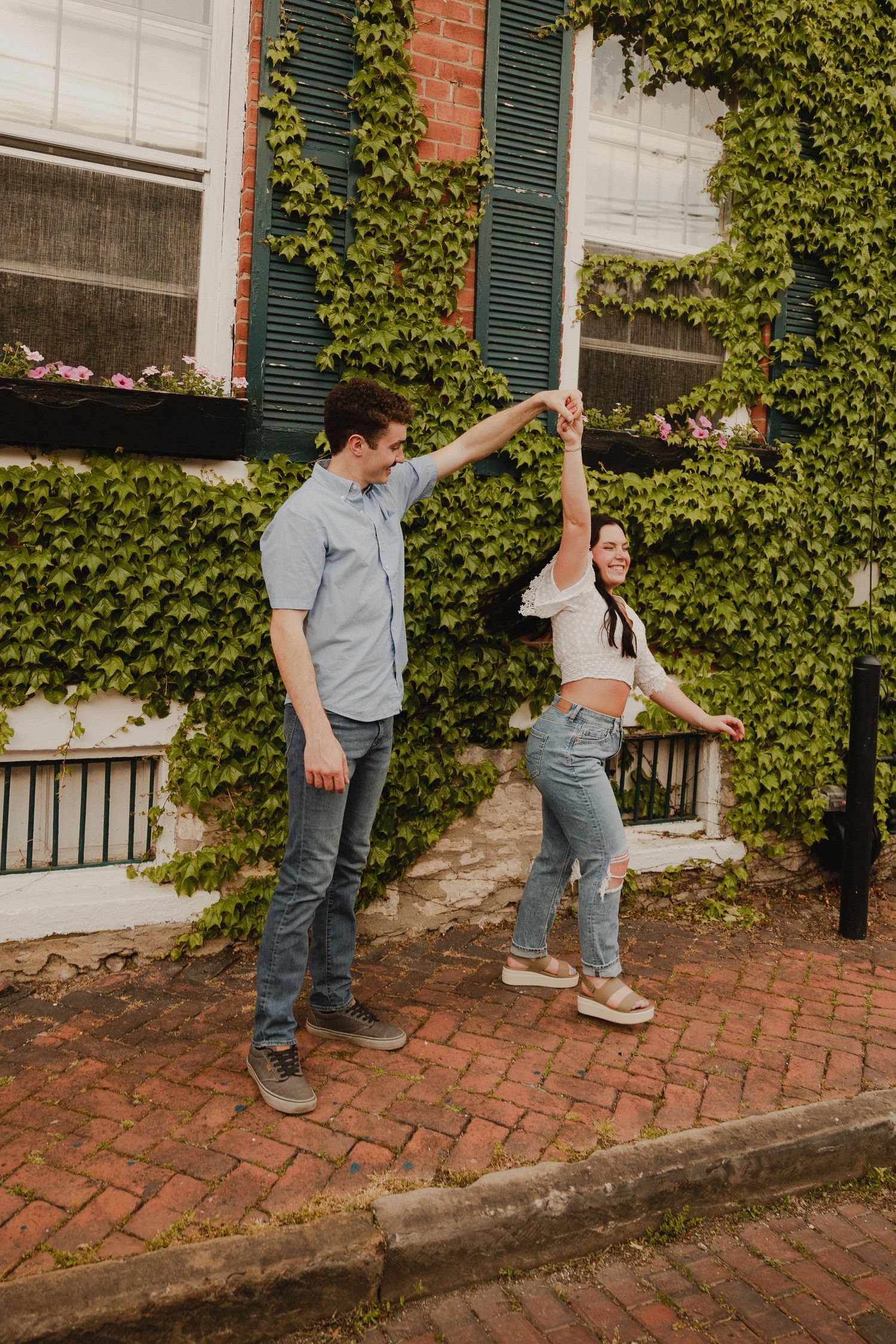 Playful dance pose on a brick sidewalk with ivy-covered building in background.