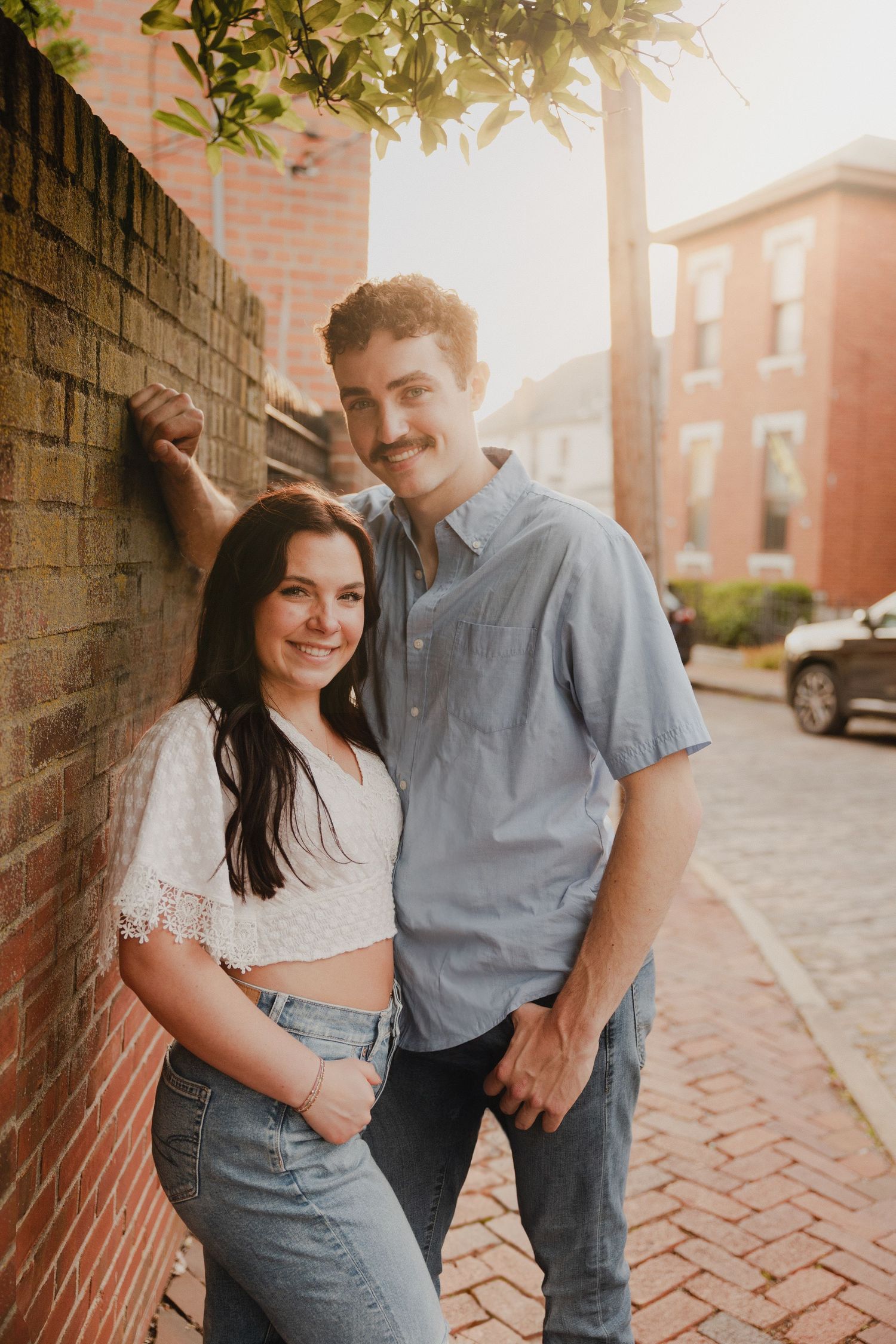 Couple posing against brick wall during golden hour on historic street.