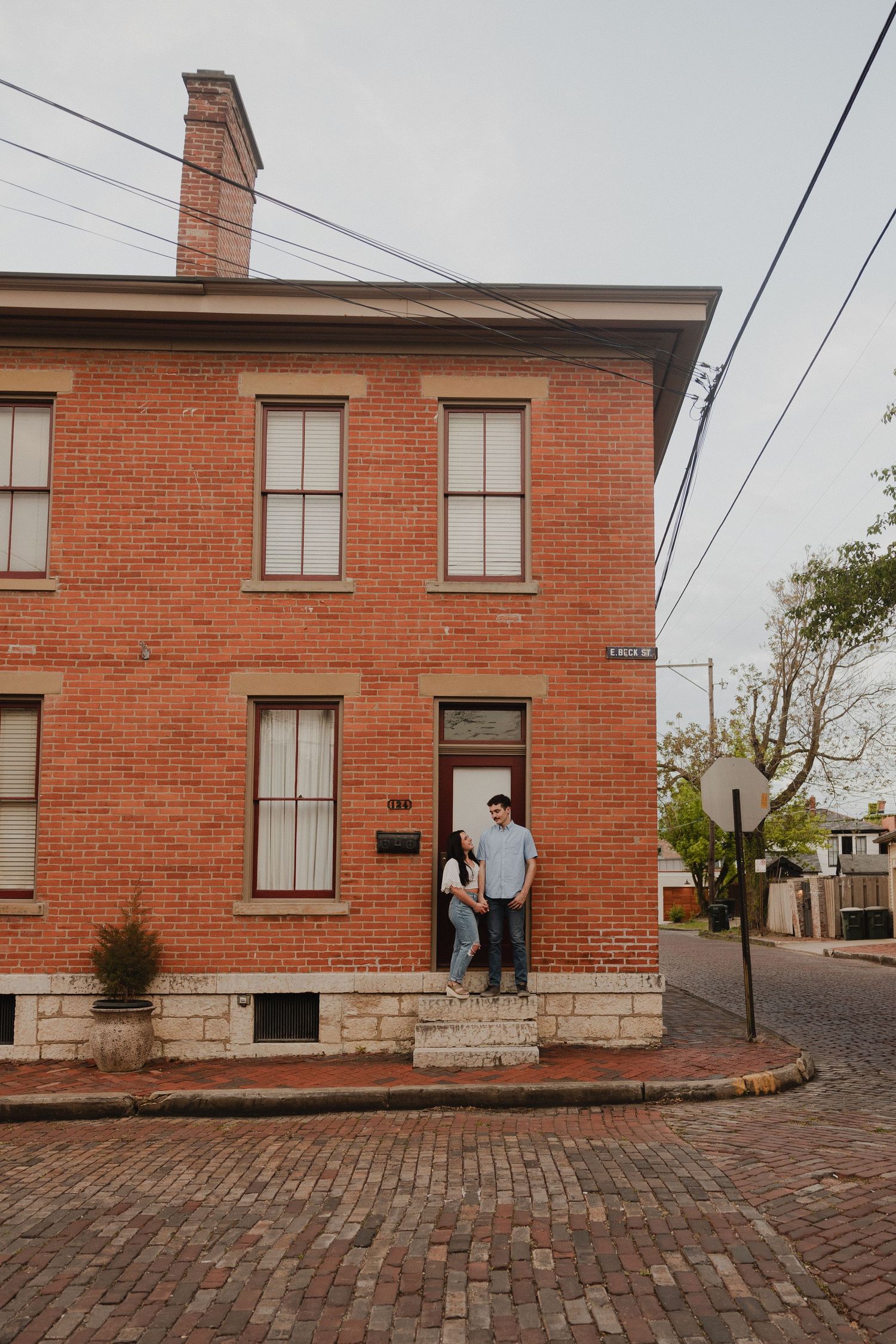 A couple stands on the steps of a historic brick building on a corner street.