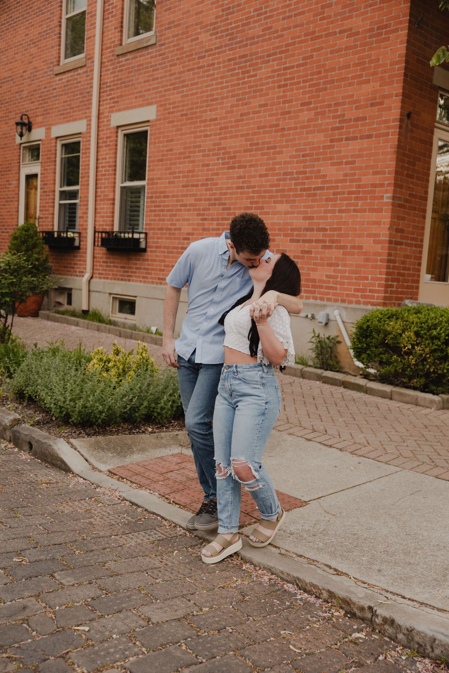 A romantic couple shares an intimate moment while standing on a brick sidewalk near a red building.