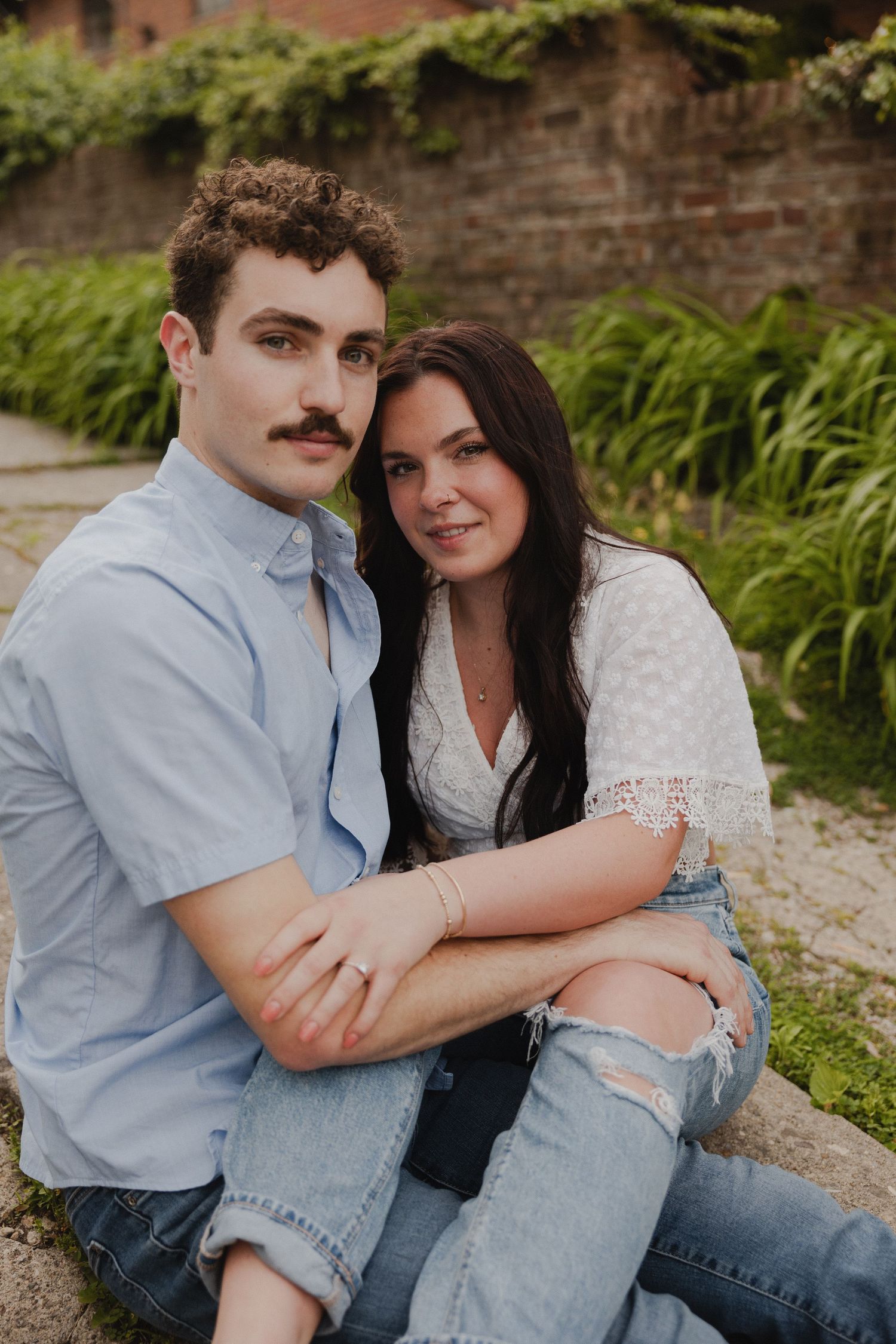 A couple in casual attire poses against a brick wall with greenery.