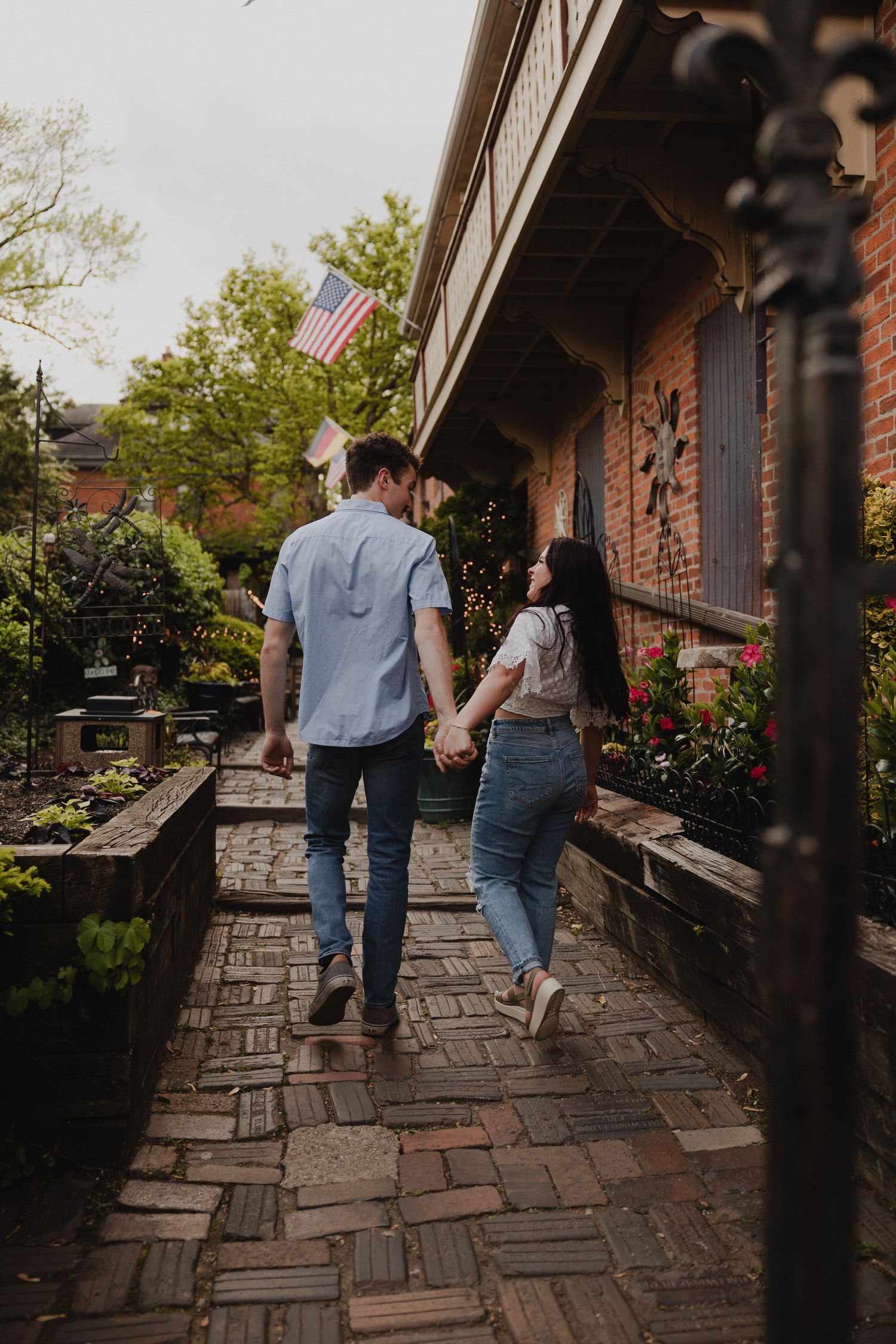 A couple holds hands while walking along a brick pathway near a garden.