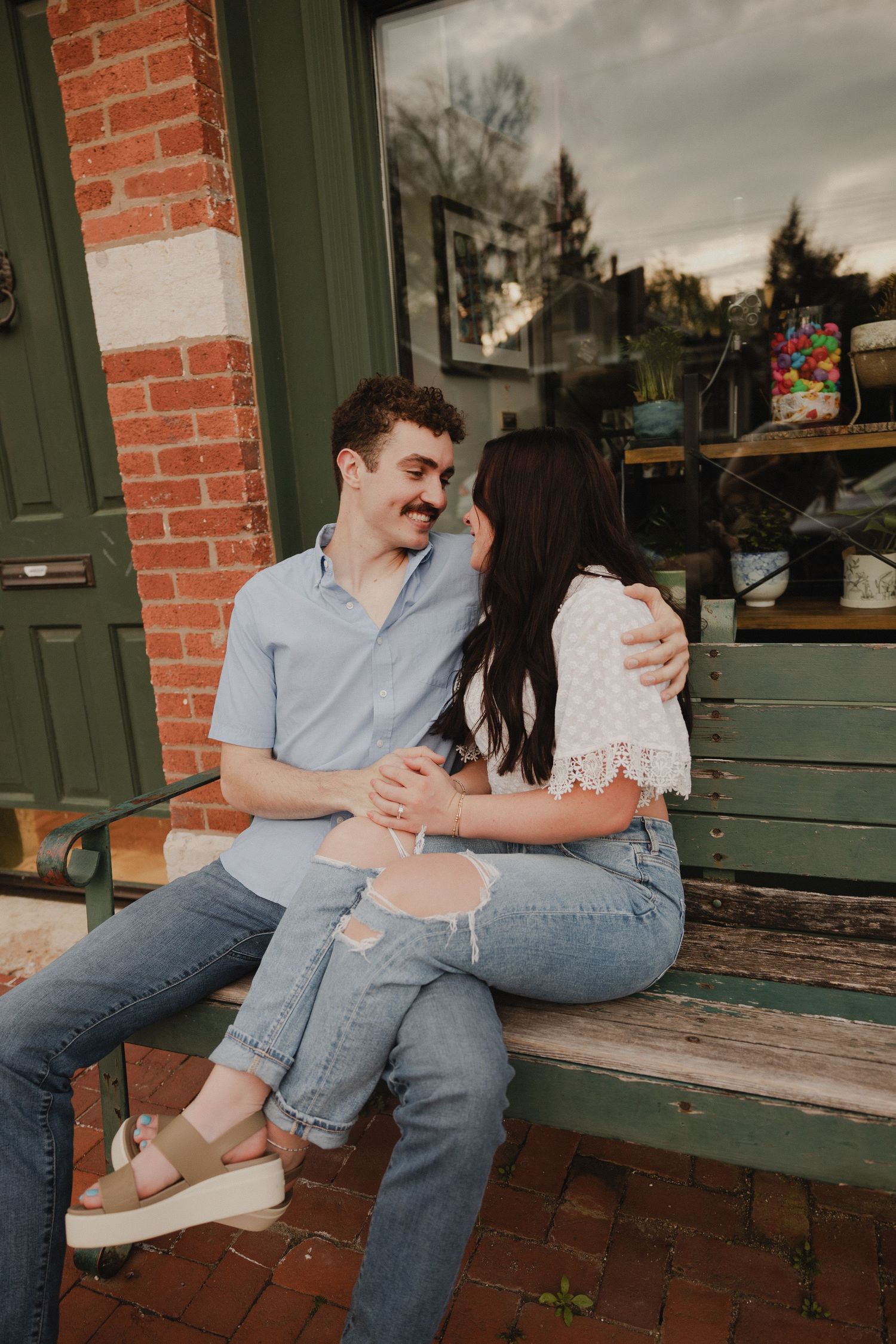 A couple shares a playful moment while sitting on a wooden bench outside a brick building.