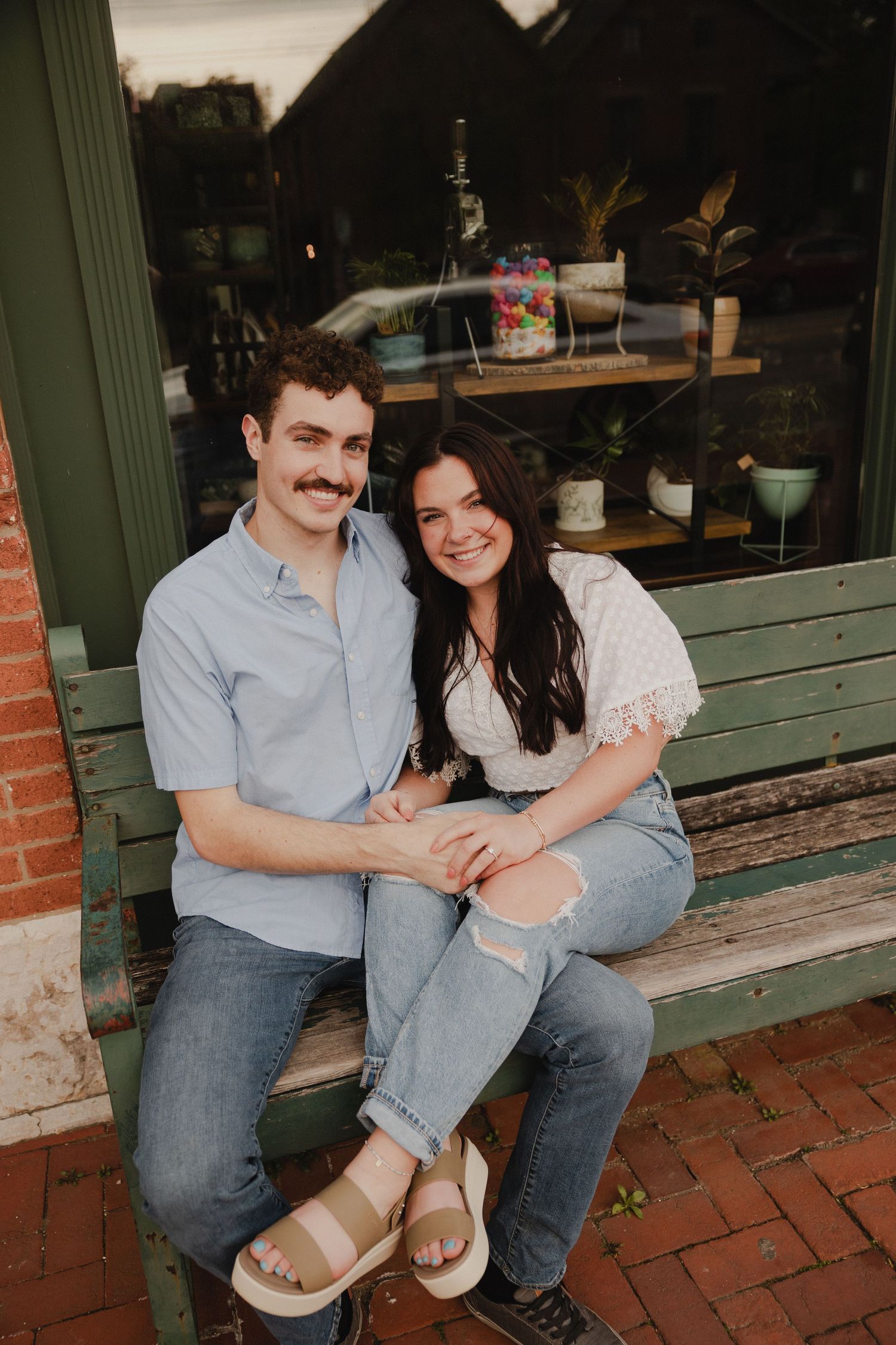 A couple poses together on a wooden bench outside a storefront.