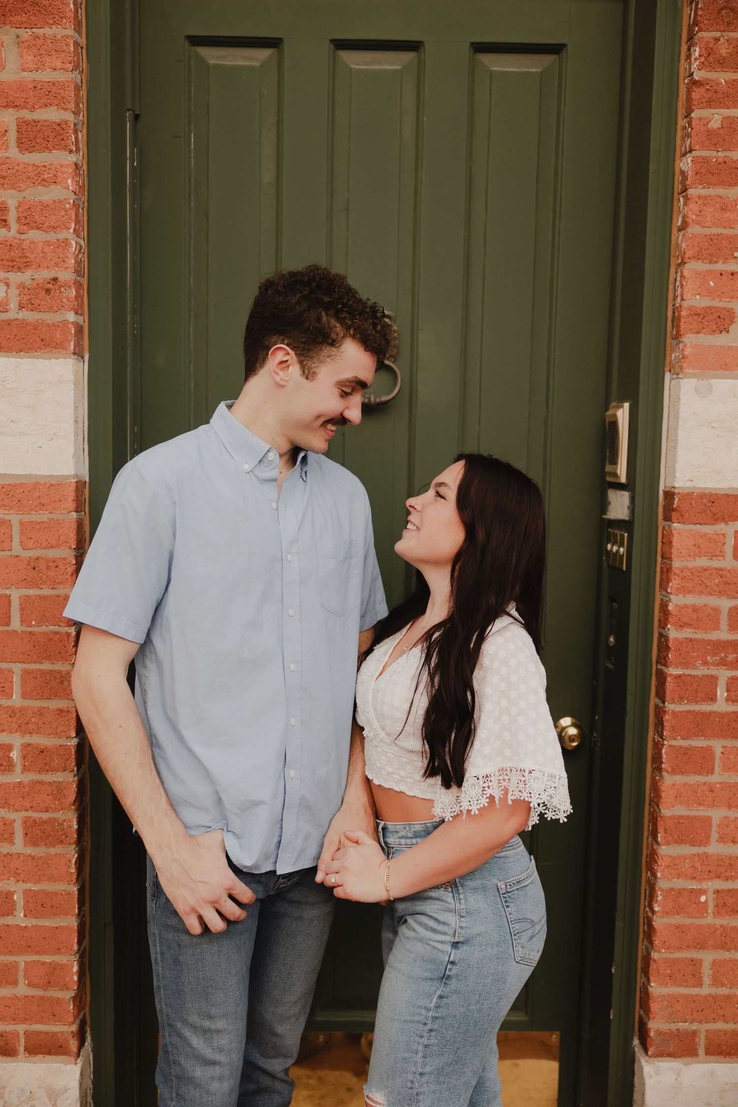 A couple shares a tender moment in front of a green door with brick surroundings.