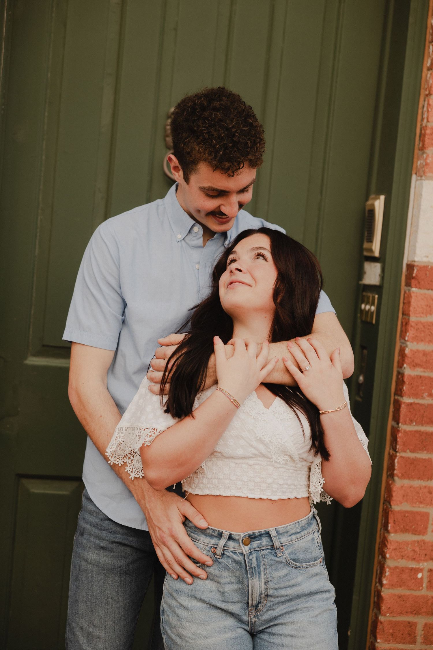 A romantic couple embraces against a green door and brick wall, with her wearing a white crop top and jeans.