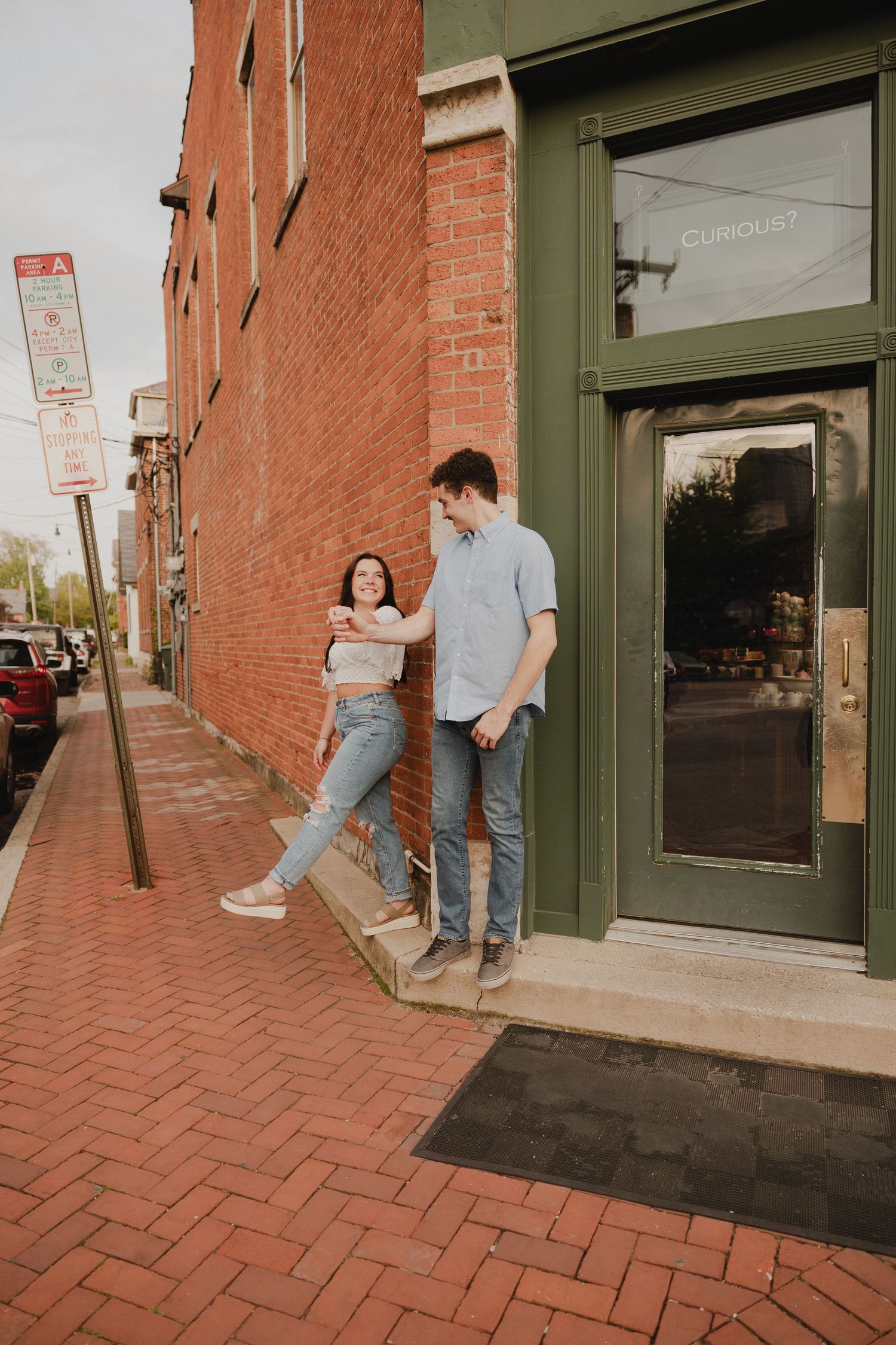 Couple poses casually against brick building with green doorway on brick sidewalk.