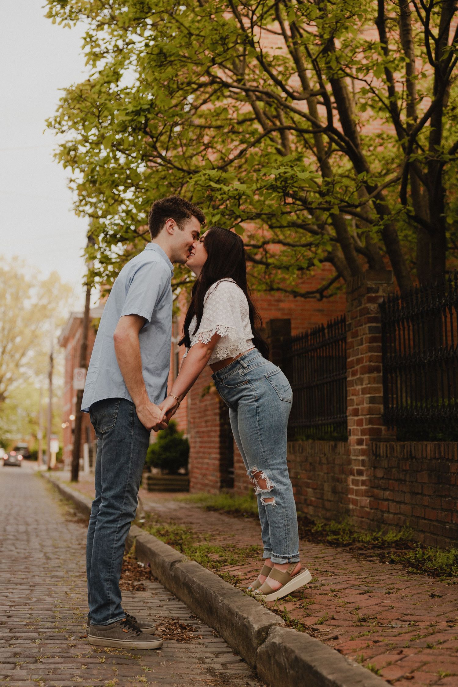 Couple shares intimate moment on cobblestone street with brick buildings and trees.