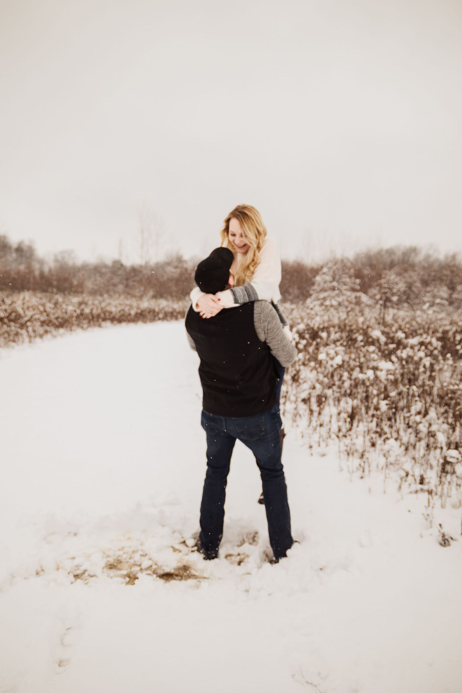 Couple embracing in a snowy field with winter vegetation in the background.