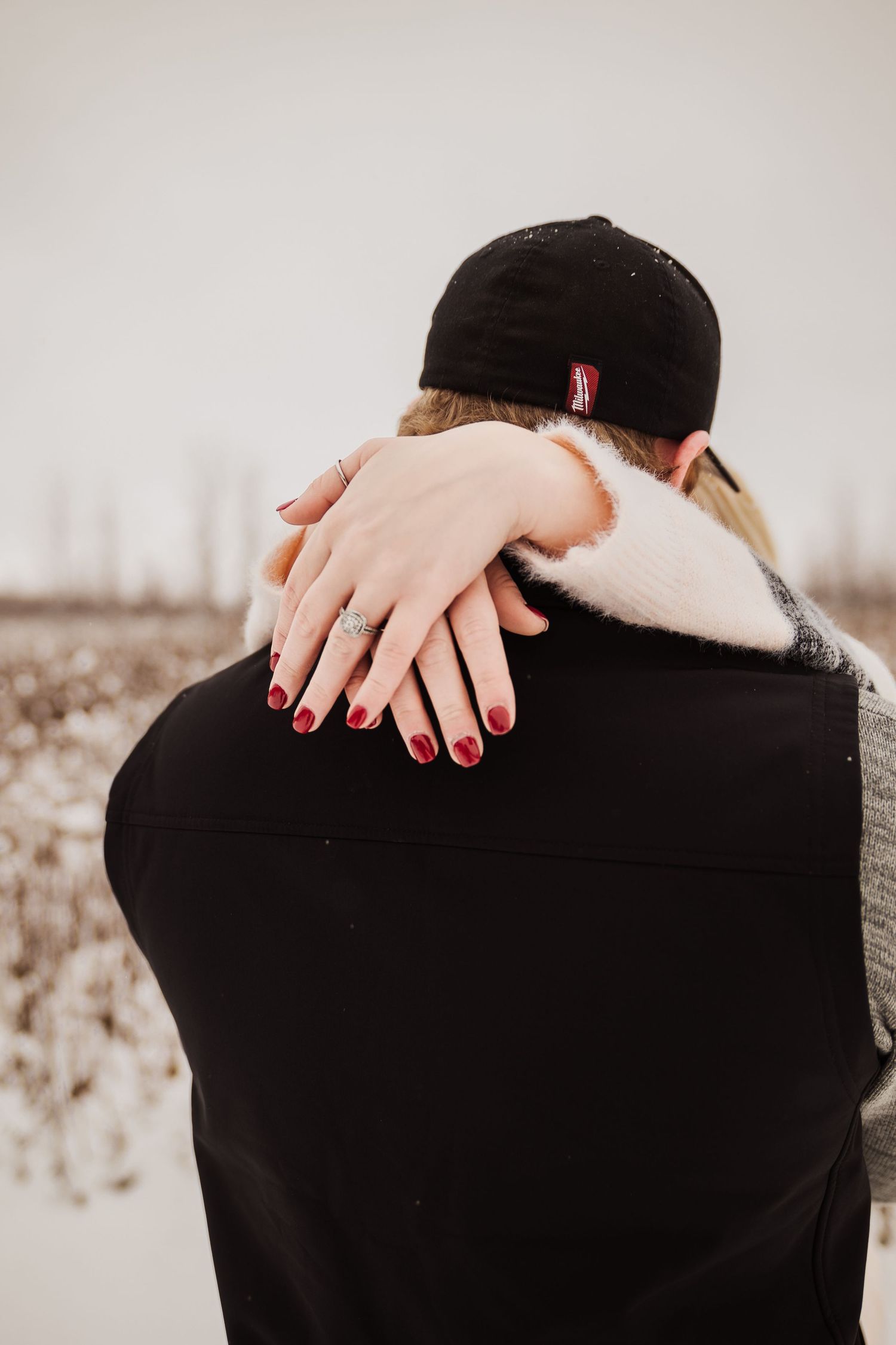 Close-up of hands embracing from behind, showing red nail polish against a black jacket.