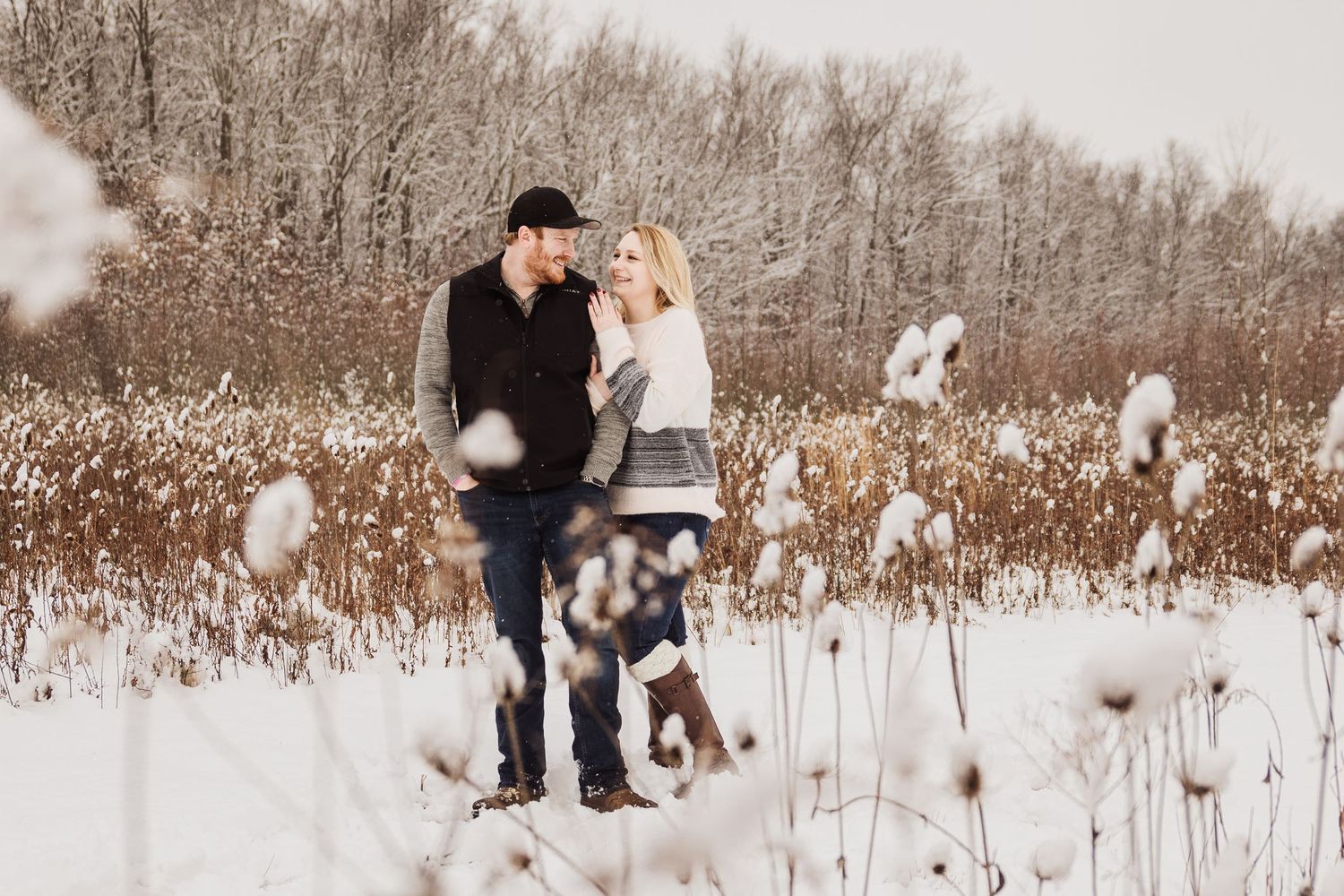 Couple standing together in a field of snow-covered cattails during winter.