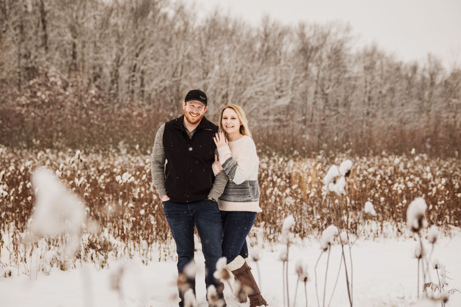 A romantic winter engagement photo of a couple walking through a snowy field with dried plants.