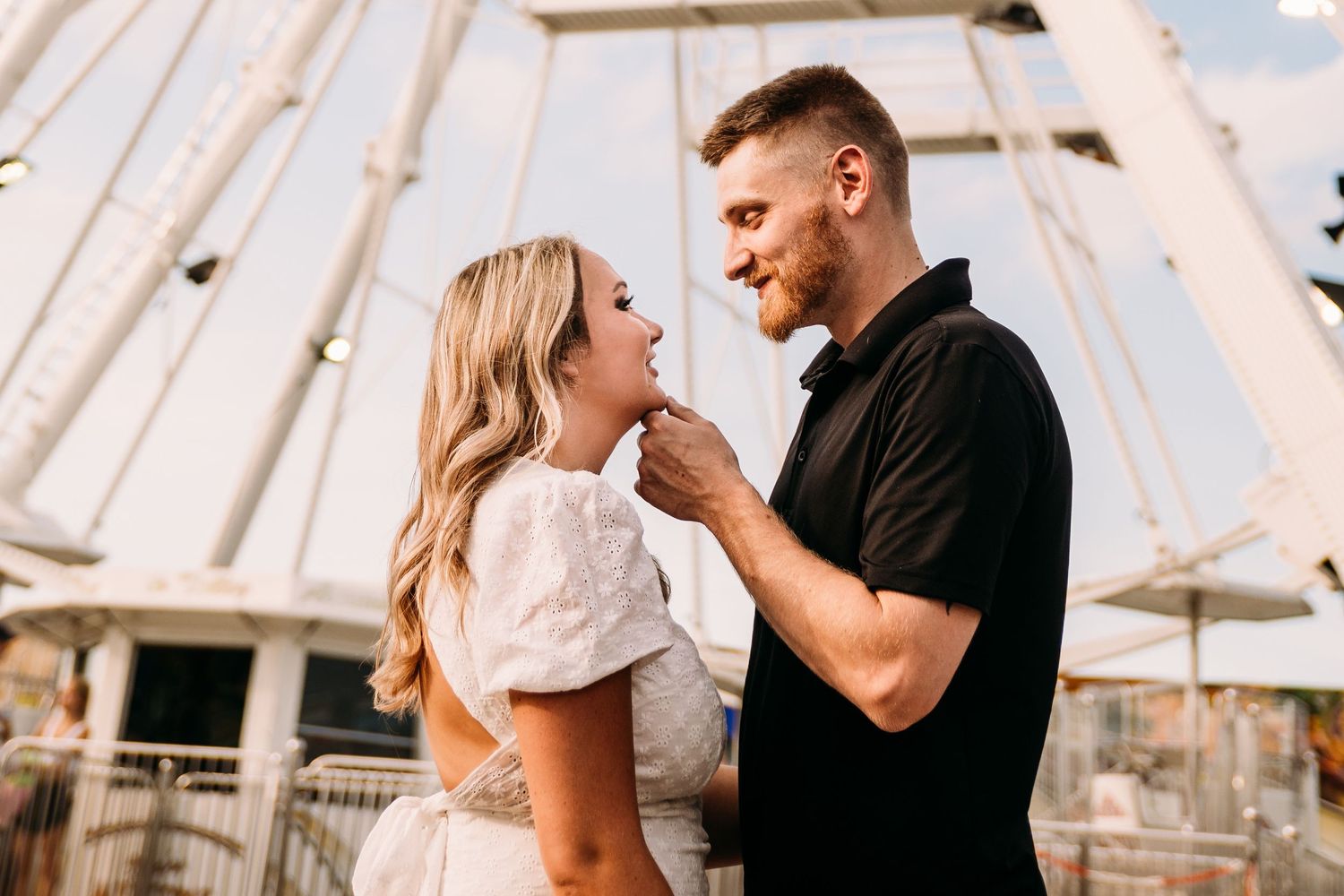 A romantic couple share an intimate moment in front of a ferris wheel at a carnival.